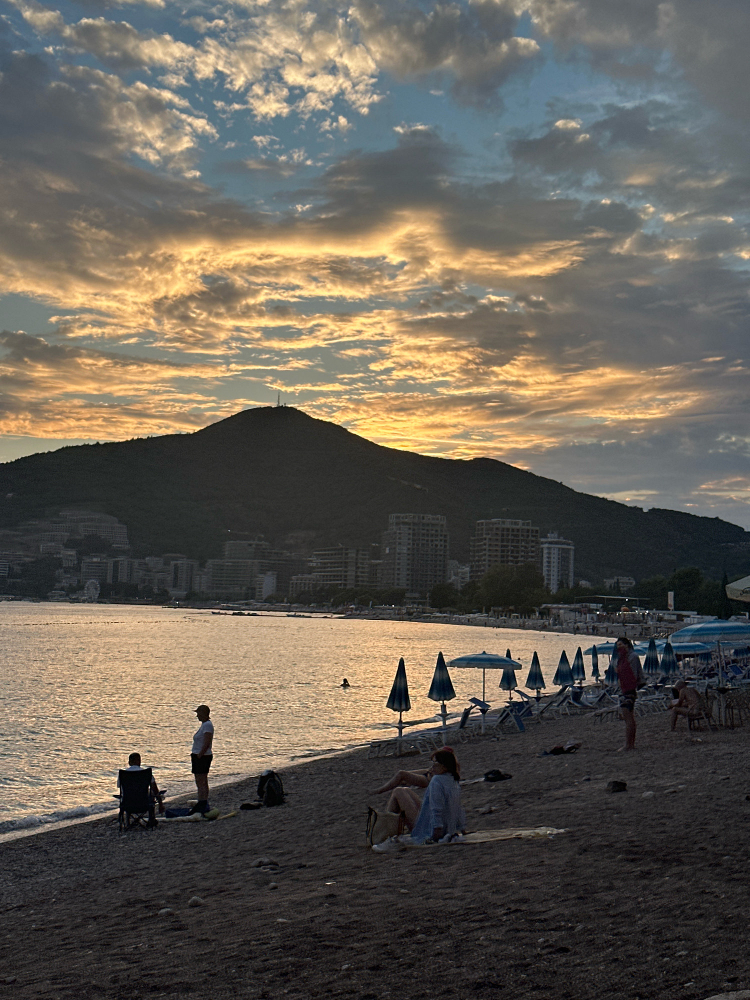 This is a photograph taken from the beach in Budva, Montenegro, at sunset. The sky is filled with dramatic clouds, some of which are illuminated by the setting sun, creating a warm, golden hue. In the background, a large, dark mountain dominates the landscape, with buildings and a transmission tower visible on its slopes. The sea is calm and reflects the colors of the sky.
On the beach in the foreground, several people are scattered. In the middle ground, there are rows of beach umbrellas and lounge chairs, suggesting a popular tourist spot. Some individuals are seen relaxing on the beach, with one person sitting in a folding chair near the water's edge. Another person stands near them. Further down the beach, more people are visible, some sitting on the sand and others standing. The overall atmosphere is serene and peaceful, capturing the beauty of a coastal evening.