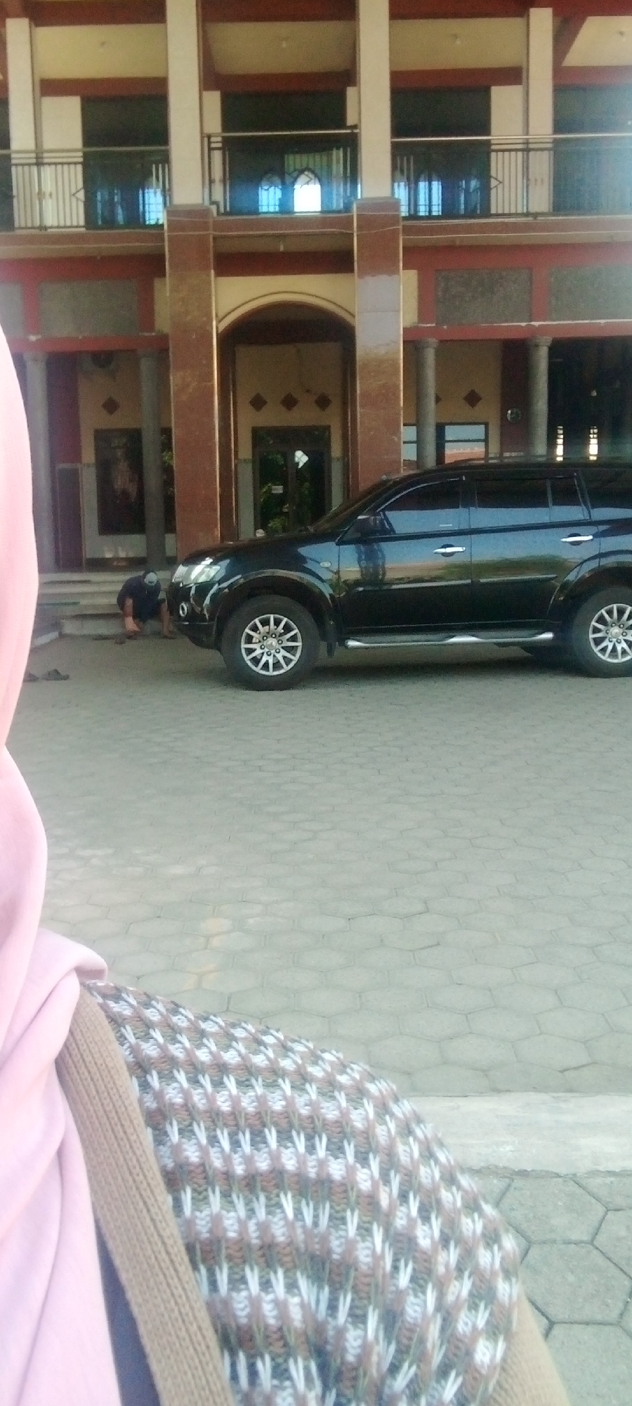 The image, captured in the city of Sumberwono, Indonesia, shows a black SUV parked on a paved surface in front of a building with a red and beige facade. The building has multiple levels with balconies and decorative pillars. In the foreground, a person wearing a pink hijab and a patterned sweater is partially visible from the shoulder up, suggesting they are the photographer or a person in the immediate vicinity. To the left of the SUV, a person wearing a dark shirt and hat is bent down, possibly interacting with something on the ground or steps leading to the building. The ground is covered with hexagonal pavers. The overall scene appears to be an outdoor urban environment, likely a public building or complex.
