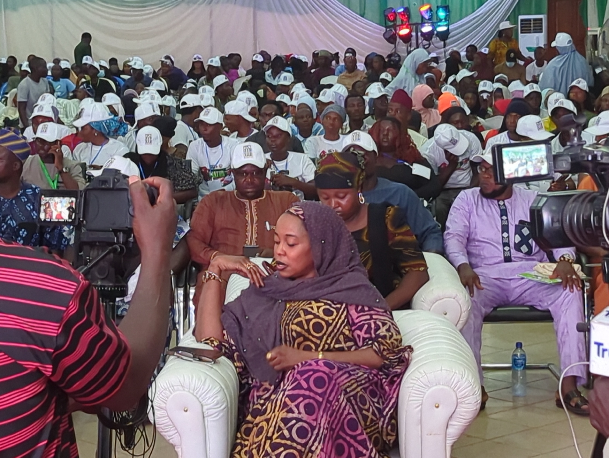 The image depicts a seated woman wearing a patterned dress and a headscarf, surrounded by a crowd of people, many of whom are wearing white caps. The scene appears to be an indoor event, possibly a conference or gathering, with stage lights visible in the background. The foreground features camera equipment and personnel. The location is Unknown, Unknown.