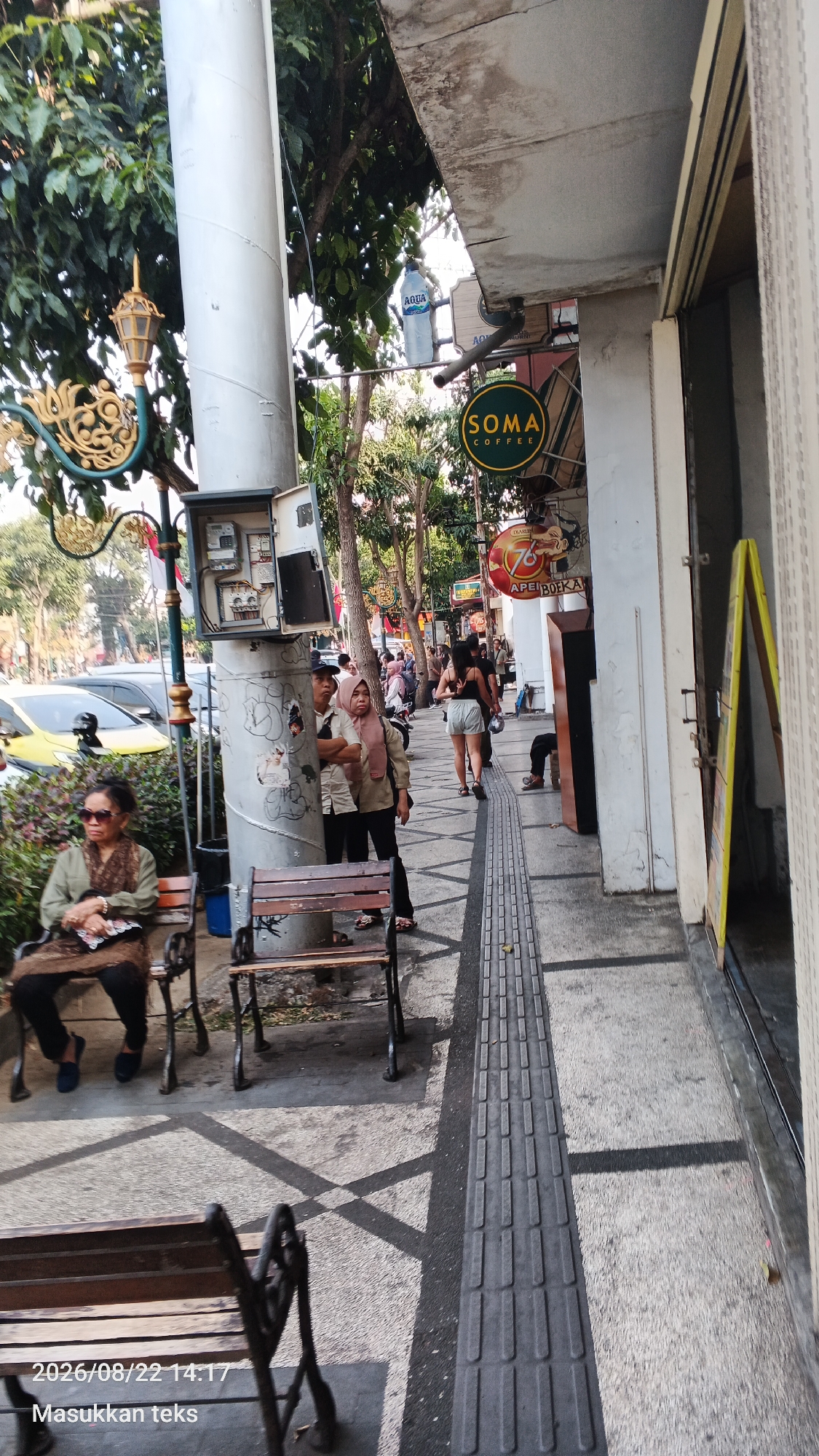 An outdoor shot captures a pedestrian walkway next to a building, likely in Malang, Indonesia. The walkway features tactile paving for the visually impaired, a common sight in urban areas. The scene depicts several individuals engaged in various activities. In the foreground, a woman sits on a bench, appearing to rest or observe her surroundings. Further down the walkway, two people stand near a utility pole, possibly waiting or conversing. A group of people are visible walking in the background, suggesting a public space with moderate foot traffic. The architecture includes elements of a traditional Indonesian building, with ornate details on a lamp post and the building facade. The presence of a "SOMA COFFEE" sign suggests a commercial establishment in the vicinity. The overall ambiance is that of a typical urban street scene in Indonesia, with elements of everyday life unfolding in a public space.