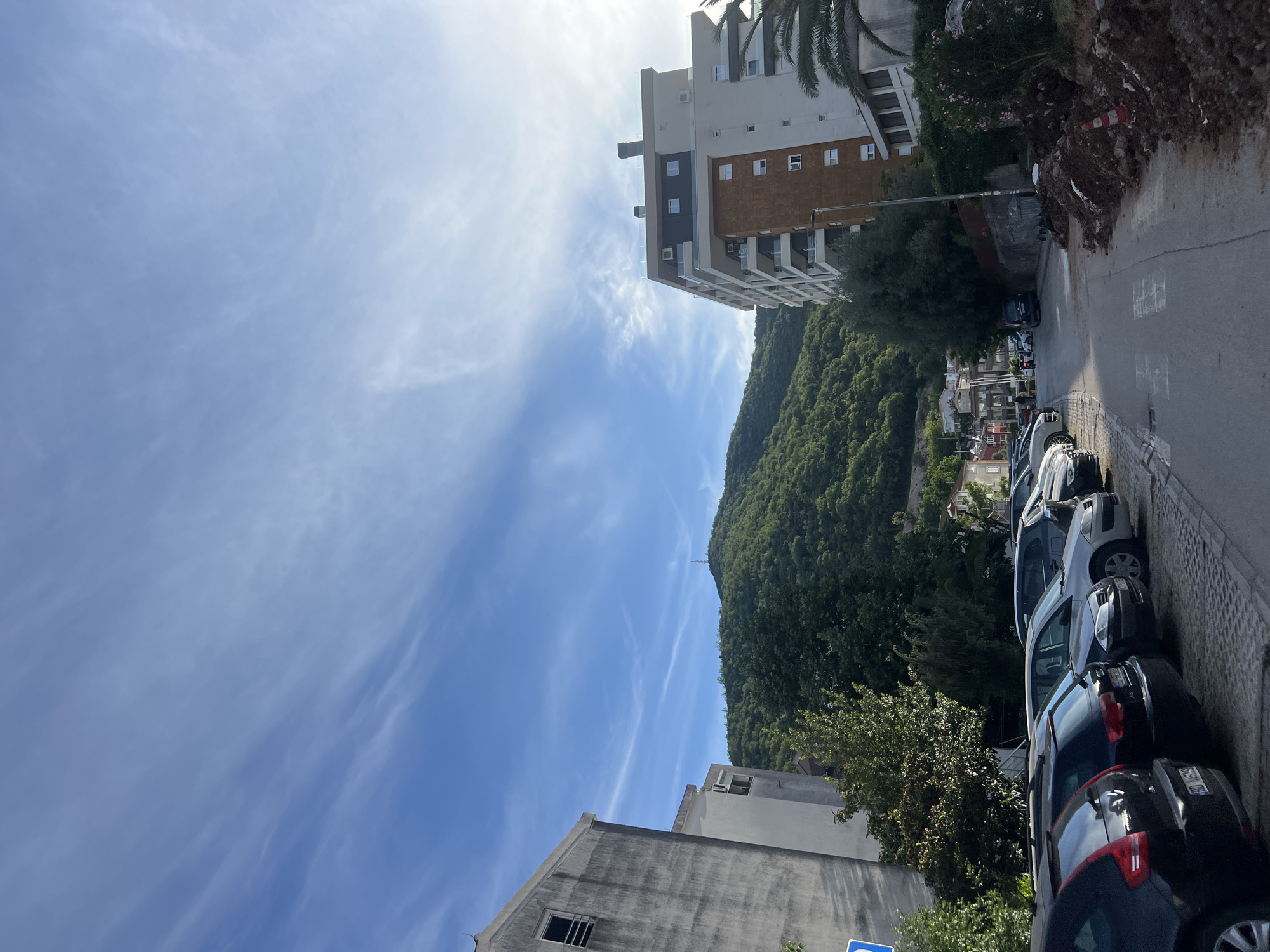 The image depicts an upward-angled street scene in Budva, Montenegro. The sky is blue with wispy clouds. On the right of the frame, cars are parked along the roadside. A building with a mix of beige and brown sections is visible above the street, along with a dense, green mountain in the background. A building is on the left of the frame, constructed of concrete. The road runs into the distance with houses and trees visible.