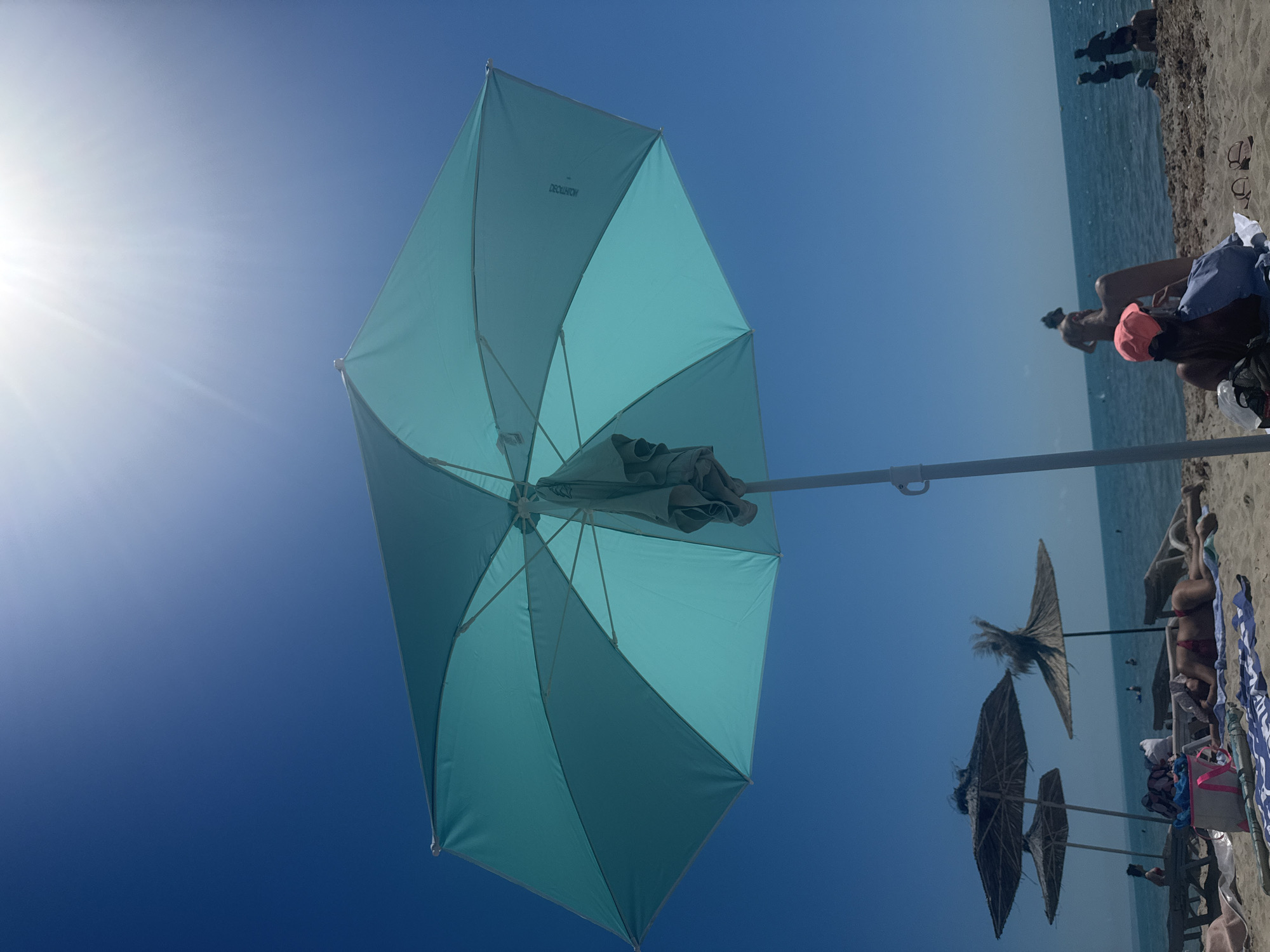 A low-angle, wide shot captures a bright, sunny day at Kranevo, Bulgaria. The sky is a clear, vibrant blue with the sun glaring intensely near the top right corner, creating lens flare. Dominating the foreground is a large, teal beach umbrella, partially open and tilted, its fabric casting shadows. A light-colored garment hangs from its central pole. Beneath the umbrella and further back on the sandy beach, several people are visible. In the mid-ground, two straw-thatched umbrellas stand out. The ocean stretches across the horizon, dotted with a few distant figures. In the foreground, a woman lies on a towel on the sand, facing away from the camera, wearing a red bikini. Another woman with a pink hat sits on the sand facing the ocean. Other beachgoers are scattered across the sand and in the water. Beach towels, bags, and sandals are present on the sand.