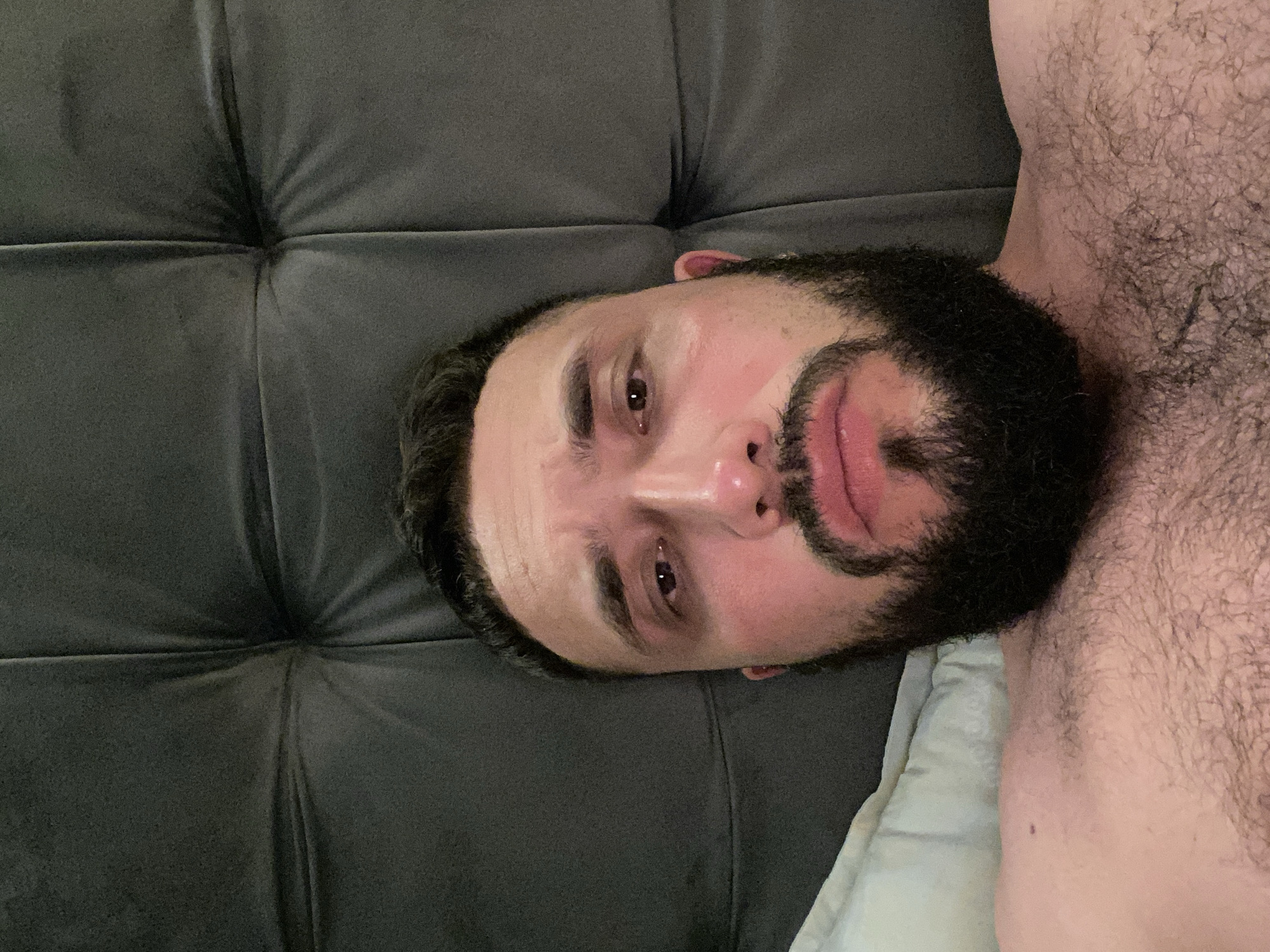 The image is a close-up, selfie-style photograph of a man with a beard and dark hair, lying down on a dark, tufted surface that appears to be a headboard or backrest. The man's head is tilted slightly and he is looking towards the camera with a neutral to slightly sad expression. The lighting suggests an indoor environment, possibly a bedroom. There is no visible indication of the city of Derbent, Russia, in the image.