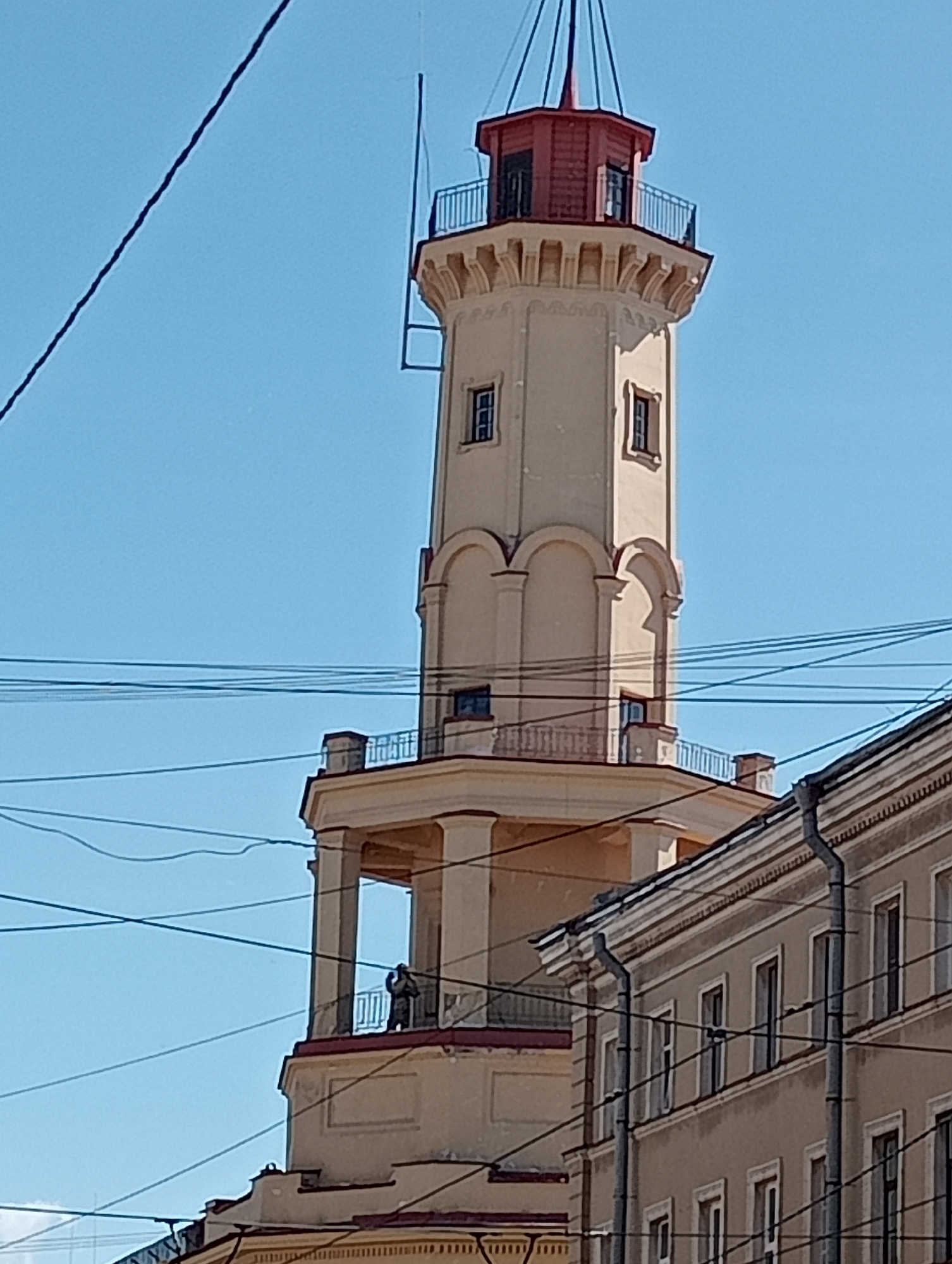 The image shows a view of the **St. Petersburg Fire Tower**, also known as the **Vigilance Tower of the Voluntary Fire Brigade**, located at **1, Sytinskaya Street, Saint Petersburg, Russia**. The tower is a tall, cream-colored structure with a red roof and a balcony. It is partially obscured by overhead wires and a neighboring building. A small, dark-colored figure, possibly a person, is visible on the balcony of the tower. The sky is clear and blue. The neighboring building is light brown with multiple windows and visible drainpipes. The overall scene depicts an urban environment under a clear sky.
