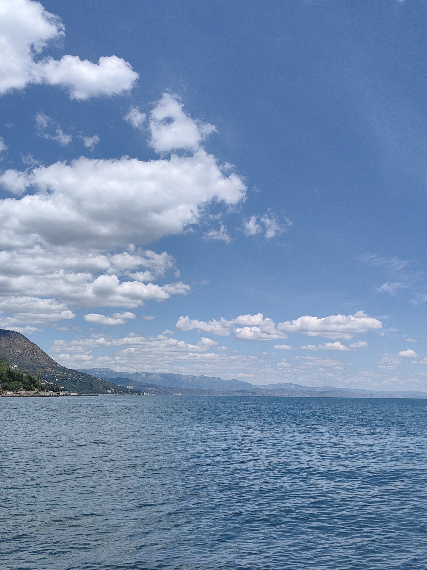 The image depicts a body of water with a coastline and mountains in the background, under a bright blue sky with scattered white clouds. The water is a deep blue and appears calm, with small ripples on its surface. The coastline is lined with trees and buildings, suggesting a populated area. In the distance, mountains form a hazy silhouette against the horizon. The scene is tranquil and appears to be a landscape view of a coastal city or town. The location is Utios, Ukraine.