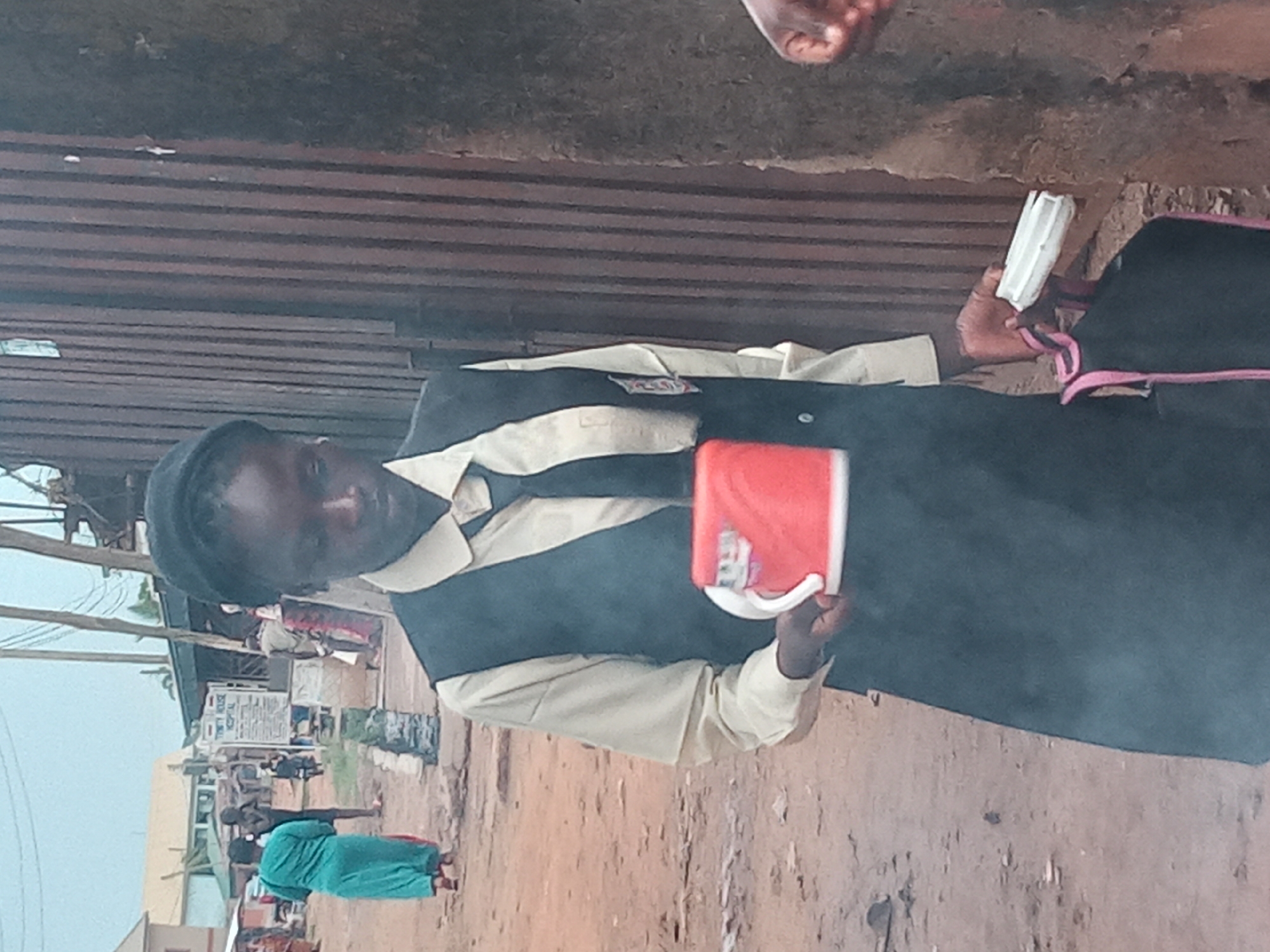 A person stands outside in Sabon Tasha, Nigeria. They wear a school uniform with a black vest over a light-colored shirt and a hat. They hold a red mug in their left hand and a white, rectangular object in their right hand. In the background, there is a building with a sign that reads "Trinity House Hospital" and other people walking around. The ground is dirt, and there is a corrugated metal structure behind the person.