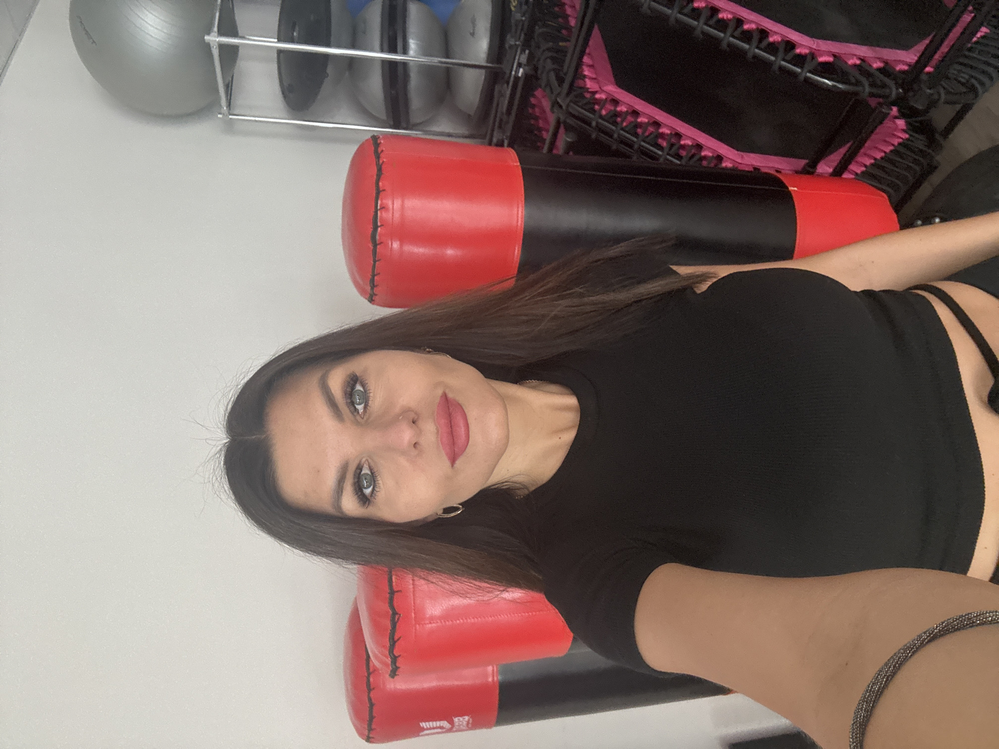 A selfie photograph shows a woman in a fitness studio setting. The woman, who has dark hair and blue eyes, is wearing a black top. In the background, there are exercise balls on a rack, a trampoline, and red-and-black boxing bags. The location appears to be in an indoor fitness area, possibly in the city of Chaikovsky, Russia.
