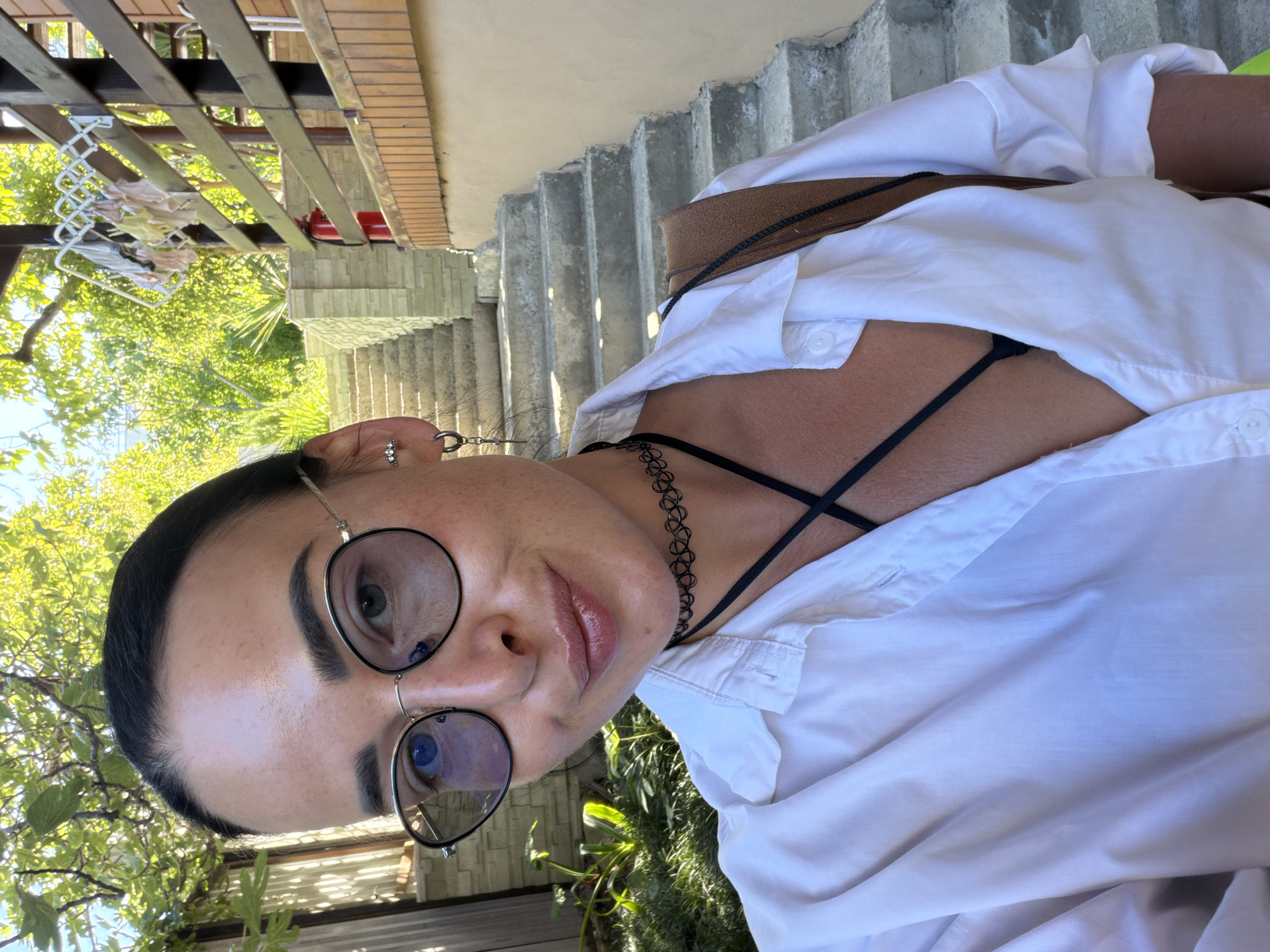 The image is a selfie of a young woman with dark hair pulled back and wearing round glasses. She is outdoors, with greenery and a stairway in the background. She is wearing a white unbuttoned shirt over a black top, and a black choker necklace. A brown bag strap is visible over her shoulder. The overall setting suggests a casual outdoor environment.