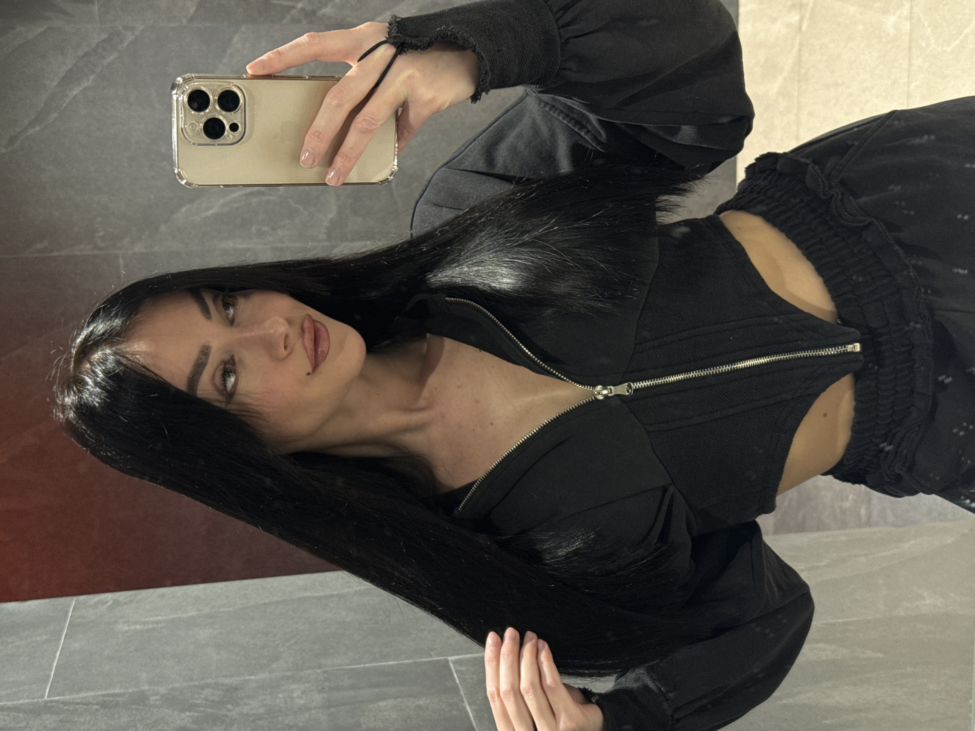 A mirror selfie captures a young woman with long, straight, dark hair, light-colored eyes, and dark eyebrows, posing with a relaxed, slightly playful expression. She is wearing a chic all-black outfit, consisting of a structured top with a prominent silver zipper running down the front and long, voluminous sleeves with distressed, frayed cuffs. A matching pair of black pants, possibly joggers, with an elasticated and ruched waistband is also visible. Her manicured nails are painted with a light, neutral polish.
She holds a light-colored iPhone, encased in a clear protective case, up to take the picture, with its triple camera array clearly visible. The setting appears to be a modern indoor space, reflected in the mirror. The background features cool-toned grey tiled walls or surfaces, with some lighter grout lines, and a darker, possibly reddish-brown, reflective surface in the upper left. The overall aesthetic is contemporary and clean. No visible text is present in the image. The location context provided is Krasnoyarsk, Russia.