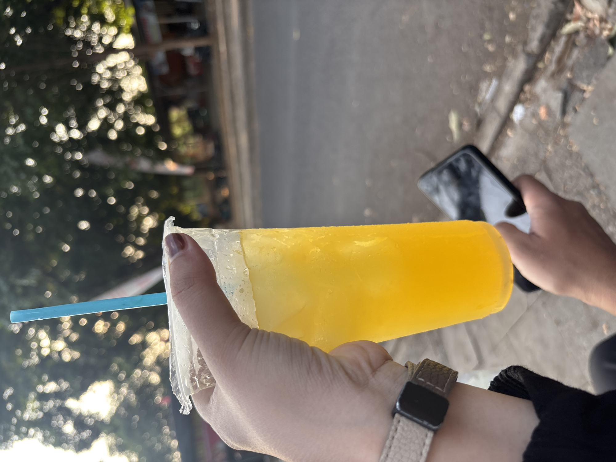 A person's hand holds a clear plastic cup filled with a yellow beverage, likely orange juice, and ice. A blue straw protrudes from the cup. The person is wearing a watch on their left wrist. In the background, another person's hand holds a black smartphone. The scene appears to be outdoors, possibly on a street or sidewalk, with trees and blurred buildings in the distance. The lighting suggests it might be late afternoon or early morning. This scene is characteristic of a street-side refreshment stall or a casual outing in the city of Cirebon, Indonesia, a common sight in bustling urban areas where vendors sell beverages to passersby.