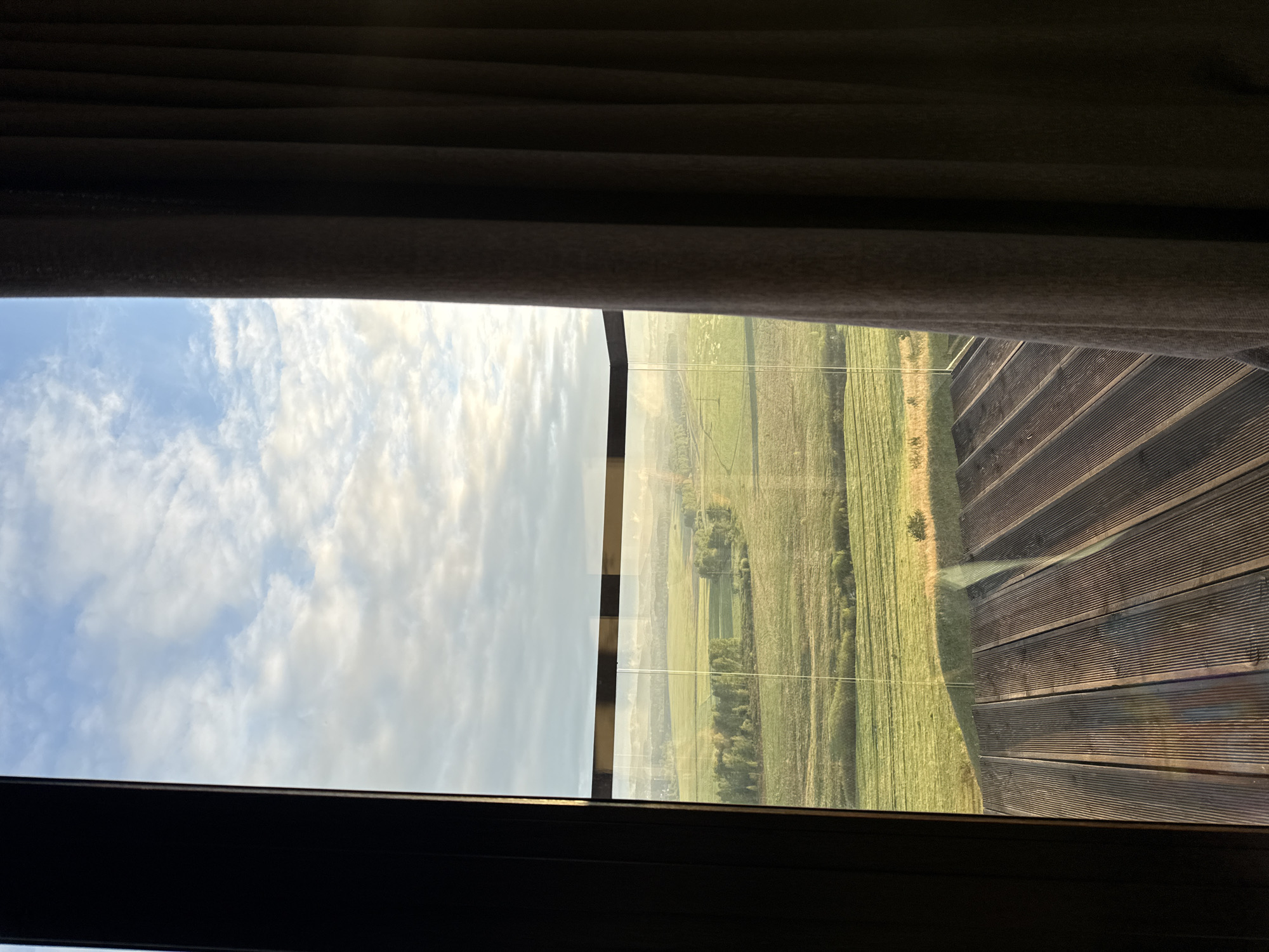 The image shows a view through a window, partially obscured by curtains. The left side of the view features a cloudy sky, while the right side shows a landscape with fields. A portion of a wooden deck or platform is visible on the right side of the window, reflecting the outside view. The scene is framed by the window's dark frame. There is no evidence to support the location of this scene being in the city of Shamoni, Russia.