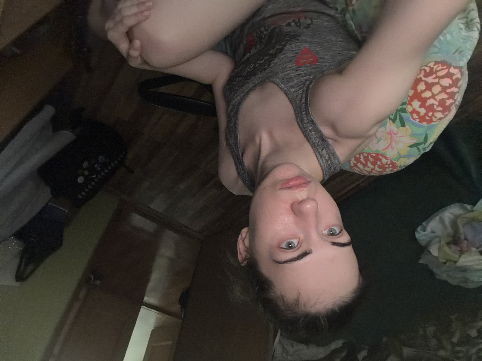 The image shows a young Caucasian female with light skin and light brown hair pulled back in a bun. She is wearing a grey tank top with a red heart design. She is seated in a chair with a colorful, pineapple-patterned cushion on the armrest. The environment appears to be a dimly lit room with wooden flooring. In the background, a doorway leads to another room, and various objects, including a dark backpack and some clothing, are visible. The woman is looking directly at the camera with a neutral expression. No specific location in Zavodoukovsk, Russia, can be identified from the visual information provided.