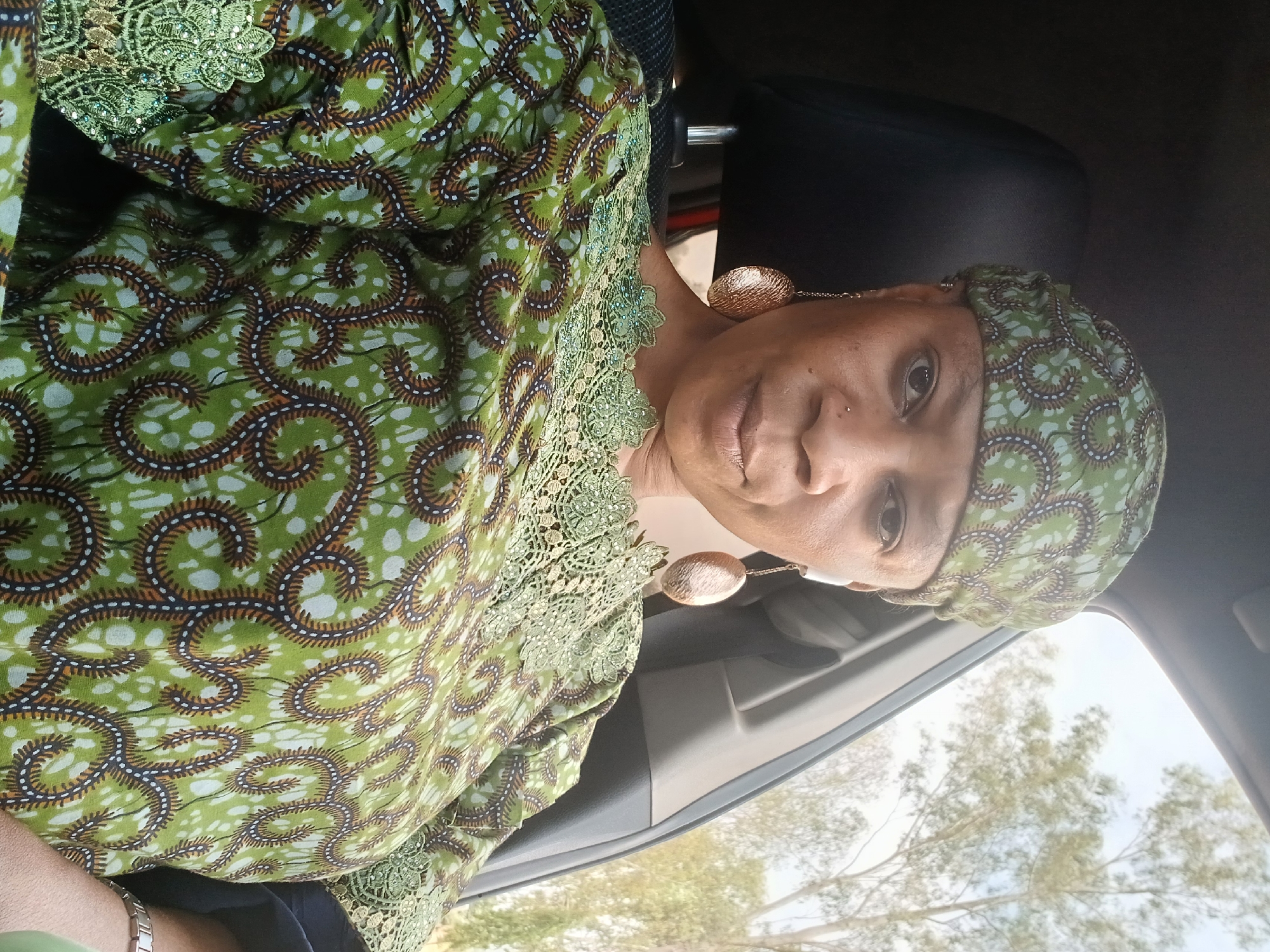 A woman is shown in a close-up shot while inside a car. She has brown eyes, dark hair, and appears to be wearing a green and brown patterned outfit with matching headwear. Large gold earrings are visible. The location is inferred to be Rigachikun, Nigeria, based on the prompt's request, but there are no distinct landmarks or features to confirm this. Outside the car window, trees and sky are visible. The woman is positioned in the driver's seat.