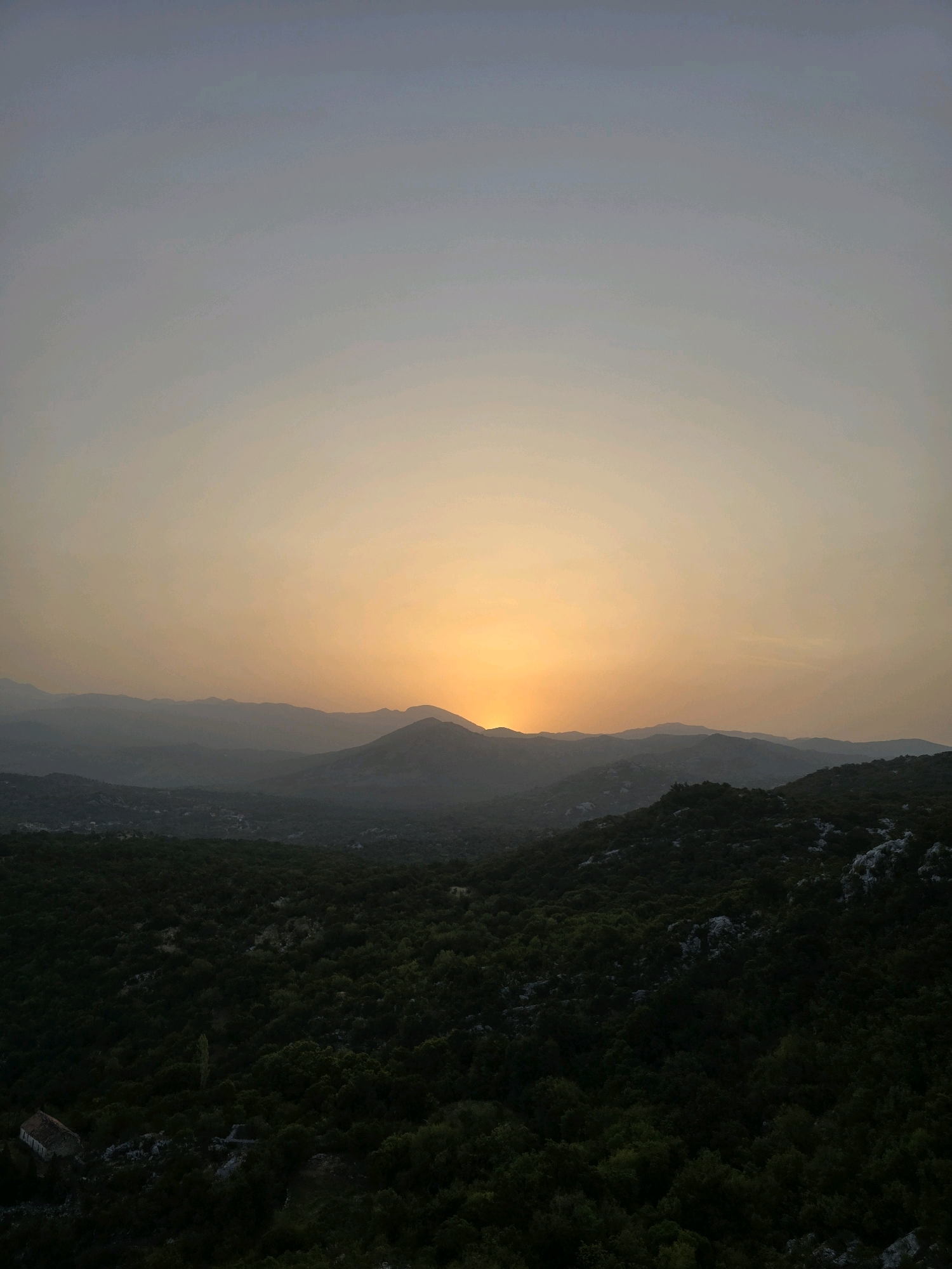 The image depicts a panoramic view of the landscape surrounding Medun, Montenegro, at dusk. The sky is a gradient of pale blue at the top, transitioning to a warm, soft orange hue near the horizon, indicating the setting sun. Layered, silhouetted mountain ranges recede into the distance, softened by atmospheric haze. The foreground is dominated by a dense, dark green forest covering rolling hills. Nestled within the trees in the lower left quadrant of the image is a small, aged building with a reddish-brown roof, appearing somewhat isolated. The overall scene is serene and natural, with no people or animals visible. The environment is characterized by undulating terrain covered in thick vegetation, with the architectural element providing a subtle human presence within the vast natural setting.