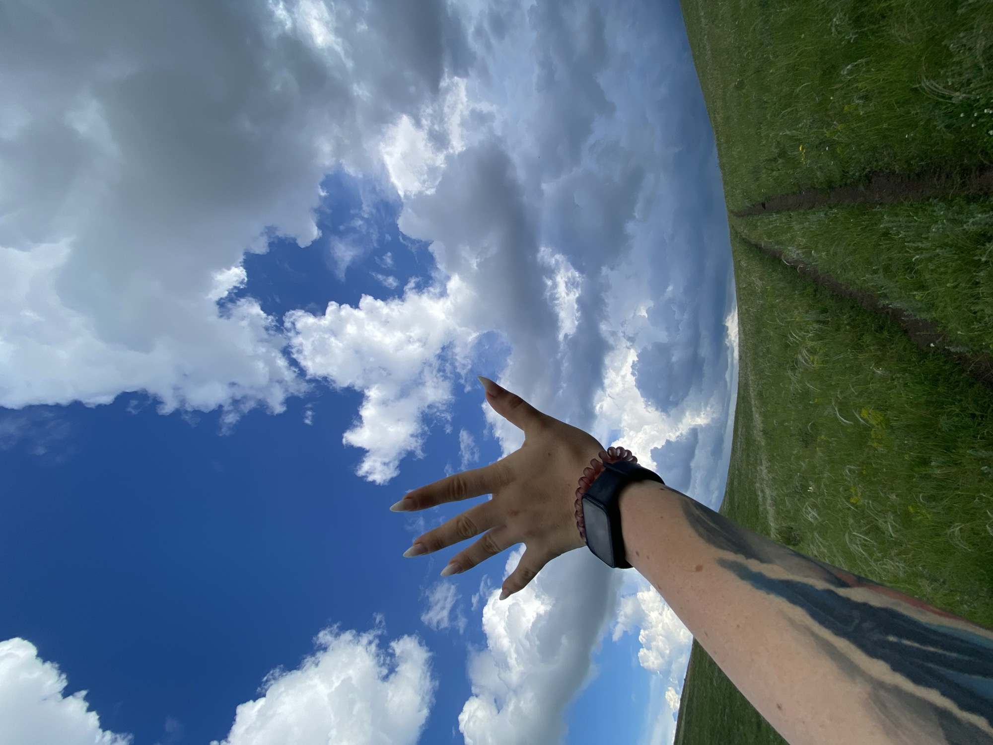 An arm with a black smartwatch and a tattoo extends into a blue sky filled with white and gray clouds. The arm is raised as if reaching for the sky. Below the sky is a rolling green hillside with a dirt path winding up it. The grass is tall and feathery, suggesting a natural, open landscape. The scene appears to be outdoors, possibly in a rural or park-like setting. There is no discernible connection to the city of Verkhneozernoe, Russia, as the image lacks any identifying landmarks or context.