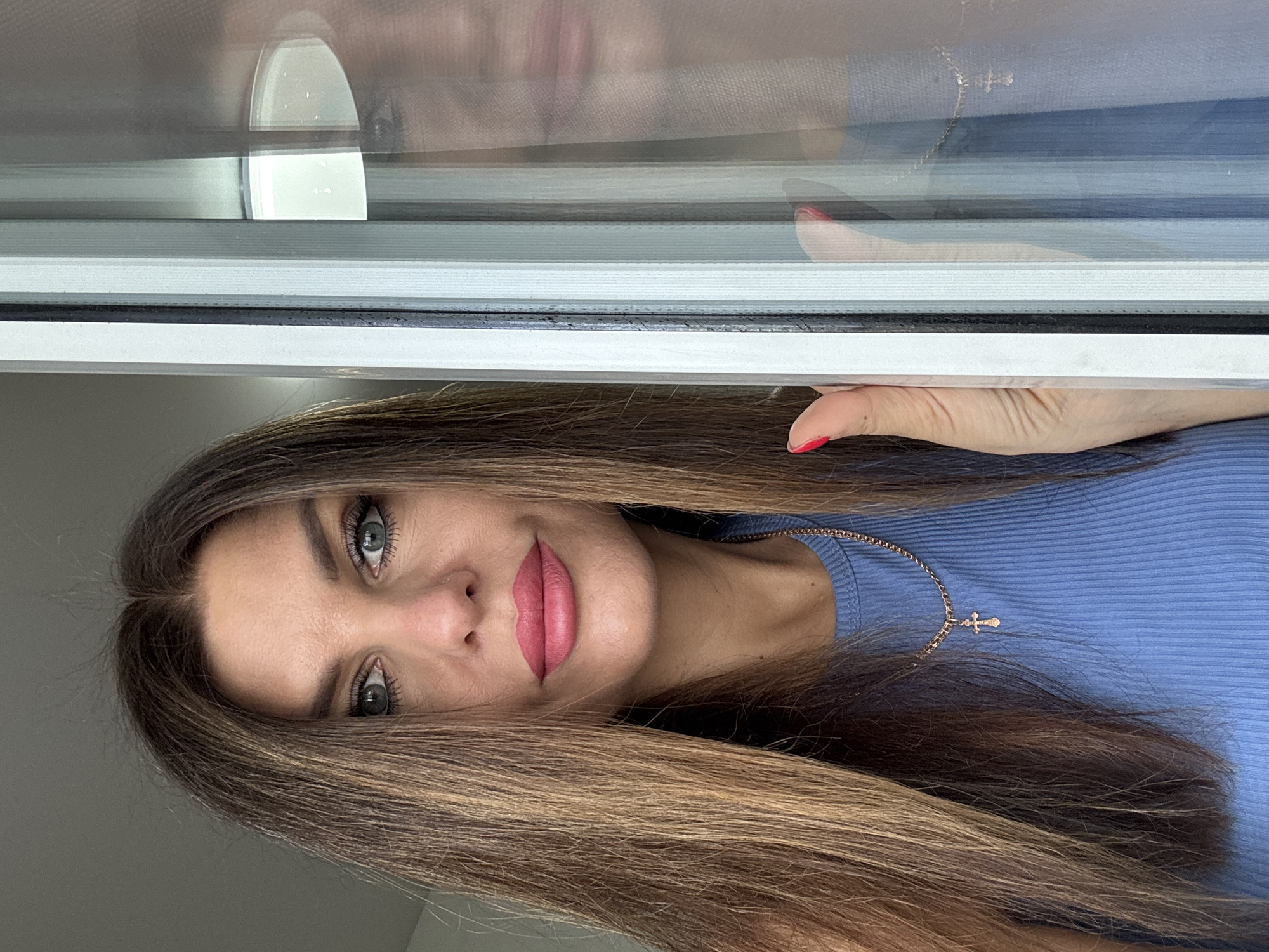 A medium shot from a slightly low angle captures a woman with long, brown, layered hair and bright blue eyes. She is wearing a blue ribbed tank top and a gold necklace with a small cross pendant. Her lips are painted a vibrant pink. She is positioned on the left side of the frame, looking directly at the camera. Her right hand is visible on the right side of the image, touching a vertical structure that appears to be a window frame or blinds. To the right of the woman, a reflection of her face and upper body is visible in the glass, slightly distorted by the vertical blinds. The background on the left is a plain, light gray wall, suggesting an indoor setting. The overall lighting is soft and diffused. There are no discernible elements in the image that indicate the specific location within the city of Chaikovsky, Russia.
