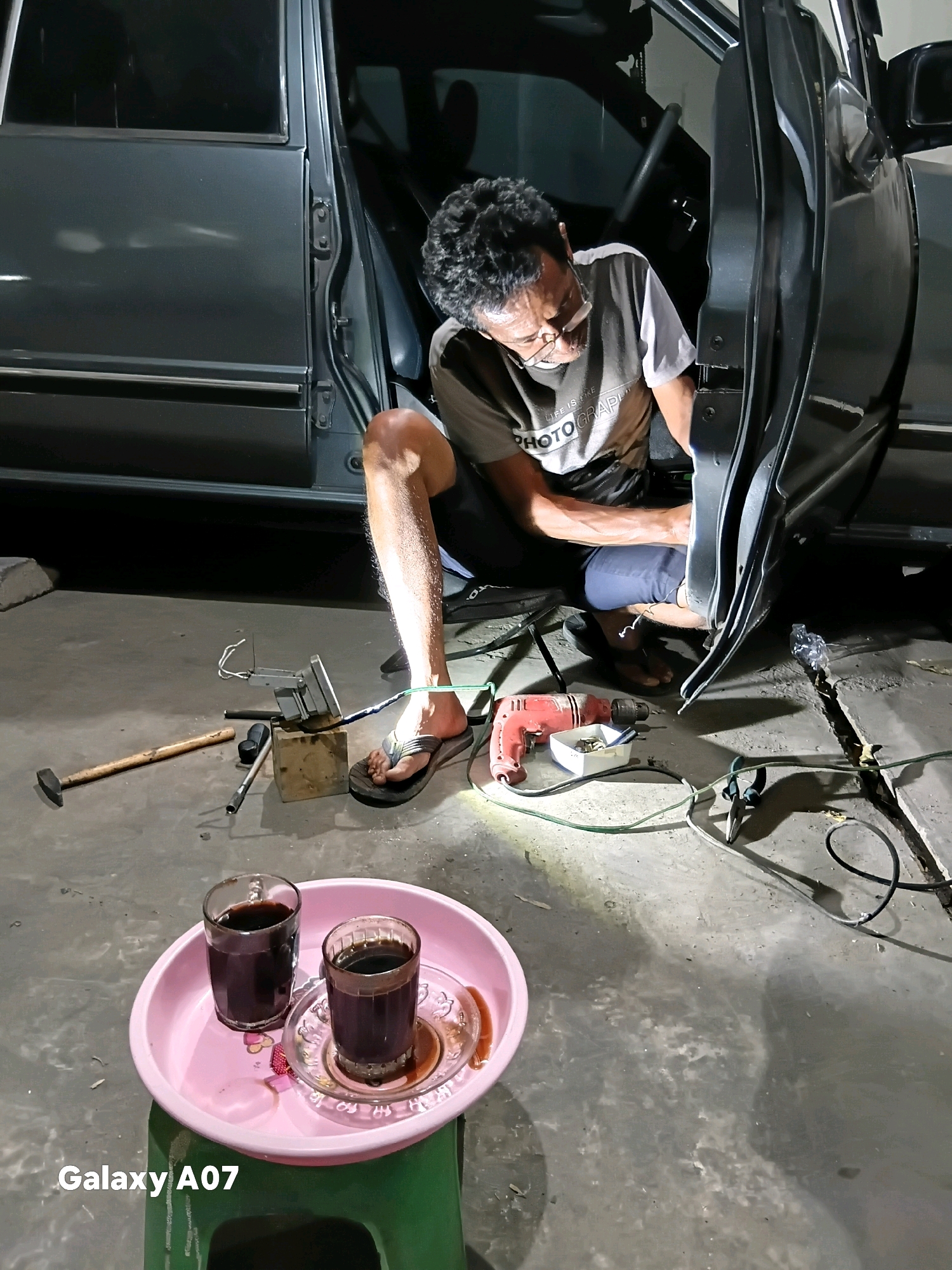 In Batu, Indonesia, a man is working on a car door, kneeling on the ground. He is wearing glasses and a t-shirt with text that reads "LIFE IS LIKE PHOTOGRAPHY." A red power drill and other tools are laid out on the concrete floor. Two glasses of dark liquid, likely coffee, are placed on a pink tray, which is on a green stool in the foreground. The car is a dark gray sedan. The scene appears to be in a workshop or garage.