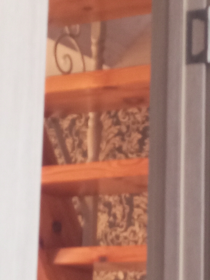The image depicts an out-of-focus, partial view of an indoor wooden staircase, framed by a light-colored vertical element on the left and a darker, possibly metallic, vertical frame with a dark rectangular attachment on the right. Multiple light brown wooden stair treads are visible, ascending towards the top right of the frame. A dark, scrolled metal decorative element, likely part of a balustrade or railing, is discernible between two upper treads. Behind the staircase, a wall or surface with a dark, intricate, and textured pattern is visible. No individuals or specific actions are present. The image provides no discernible features or geographical markers to confirm the location as the city of Яромаска, Russia.