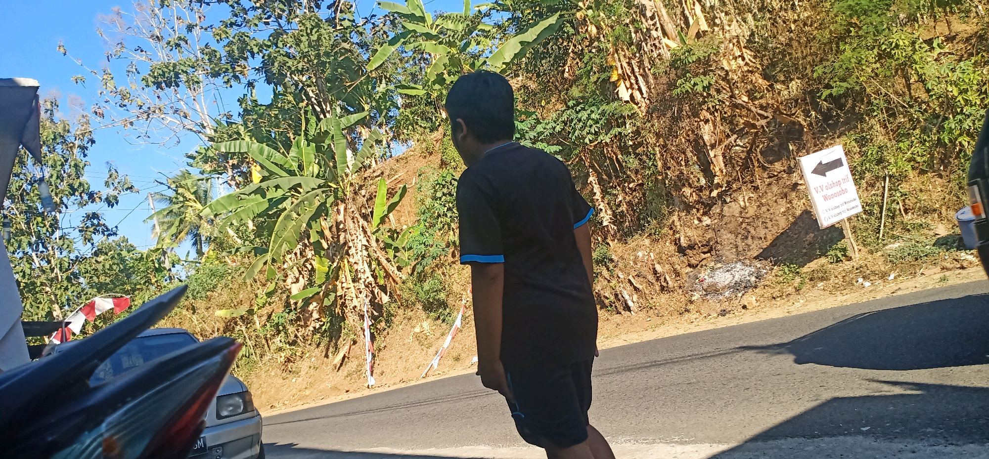 The image depicts a sunny outdoor scene with a road and a vegetated hillside. In the foreground, a person wearing a black t-shirt and shorts stands with their back to the camera, facing the hillside. To the left, parts of a vehicle, possibly a motorcycle, are visible, including handlebars and what appears to be a fender. A red and white striped flag is also partially visible. The hillside is covered in lush vegetation, including banana trees and other plants, and appears to be somewhat overgrown. A white signpost with a black arrow pointing to the left is planted on the hillside, bearing text that reads "V.V. olshop trif Wonosobo". Further down the road, a silver car is parked. The sky is clear and blue. The overall impression is a roadside scene in a rural or semi-rural area. The sign suggests the location is near Gambarsari, Indonesia, with a destination indicated as Wonosobo.