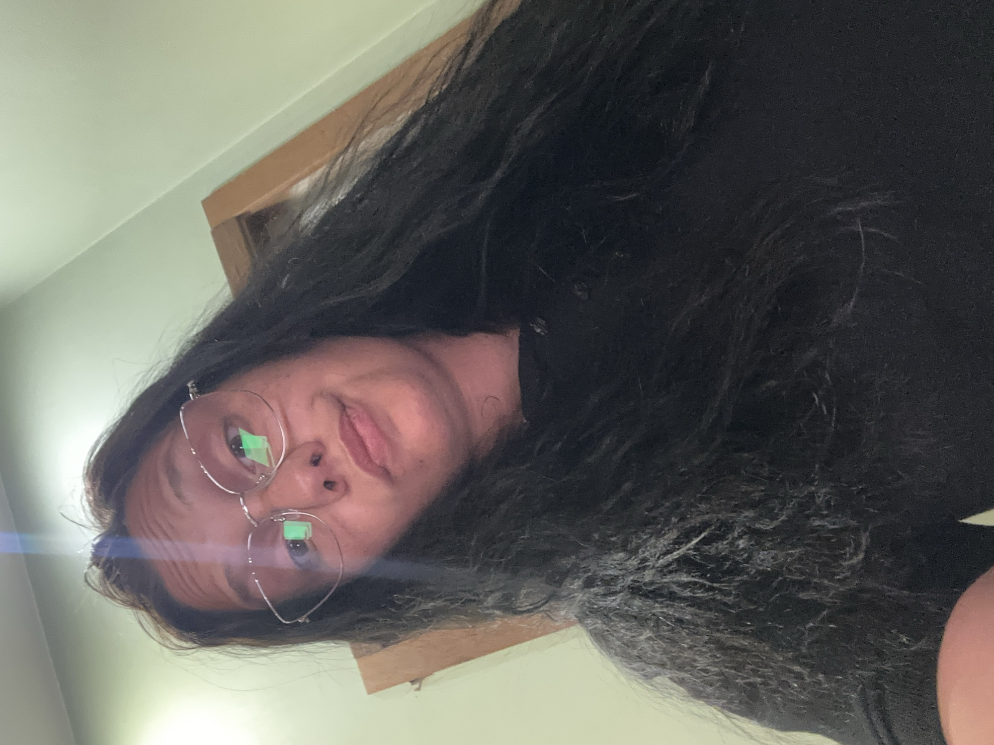 The image is a low-angle selfie of a woman with dark hair and glasses. Light is reflecting off the lenses. She is wearing a black shirt. The background shows a light green wall and a wooden frame. It's impossible to determine the location in the city of Sofia, Bulgaria from this image.