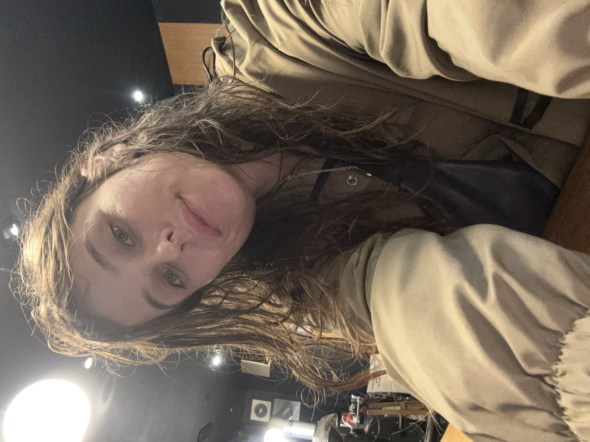 The media file is a close-up, low-angle selfie of a person with long, brown, wavy hair. The person is wearing a tan jacket with the hood up. The background is dark with some overhead lighting and what appear to be various pieces of equipment. The lighting suggests it is likely nighttime or indoors. The person's eyes are open and looking towards the camera, and their mouth is slightly parted. They appear to be in a casual setting, possibly a studio or workshop given the visible equipment. There is no visible text in the image. The location context provided is Seogwipo-si, South Korea.