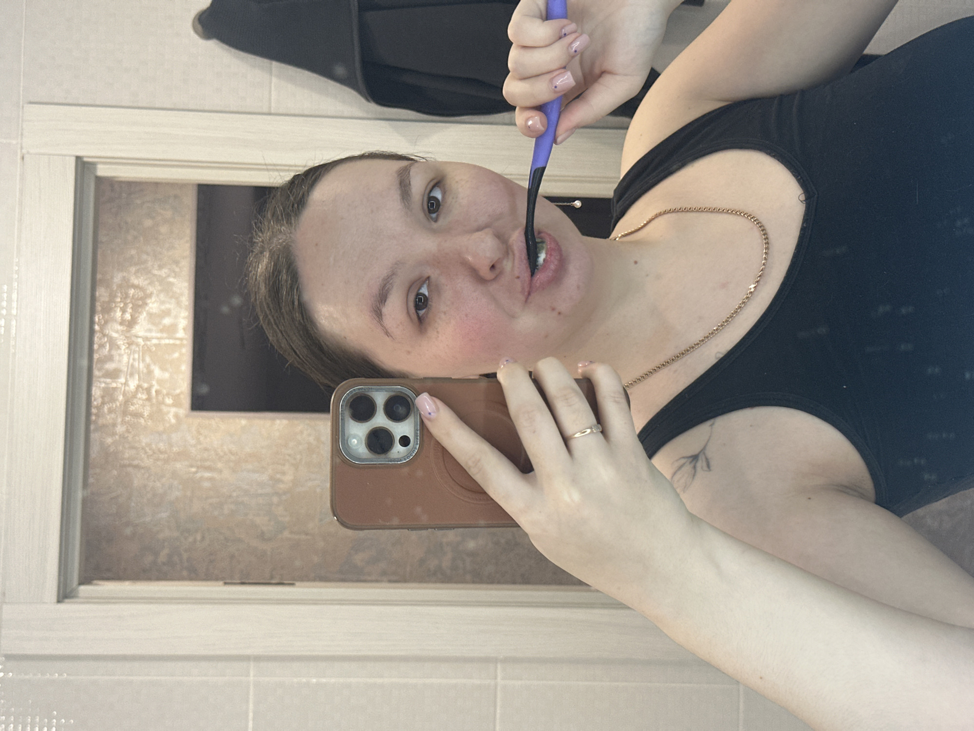 This image captures an adult female taking a mirror selfie in what appears to be a bathroom located in the city of Chaikovsky, Russia. The female, with fair skin and dark hair, is depicted from the upper chest upward, actively brushing her teeth. She holds a purple-handled, black-headed toothbrush in her right hand, with white toothpaste foam visible in her mouth and on the bristles. Her left hand holds a brown-cased smartphone (identified as an iPhone 14 Pro Max by its distinct triple-lens camera module) extended towards the mirror, capturing the image. She wears a black V-neck tank top, a thin gold chain necklace, and a simple gold band on the ring finger of her left hand. Her fingernails are manicured with light pink polish featuring small purple dots. A faint, fine-line floral tattoo is visible on her upper left arm/shoulder area. The background elements visible in the reflection include light beige tiled walls to the left, a light-colored door frame, and a reflection of a textured brown surface visible through an open doorway. A black towel hangs on a hook or rack to the right of the frame.