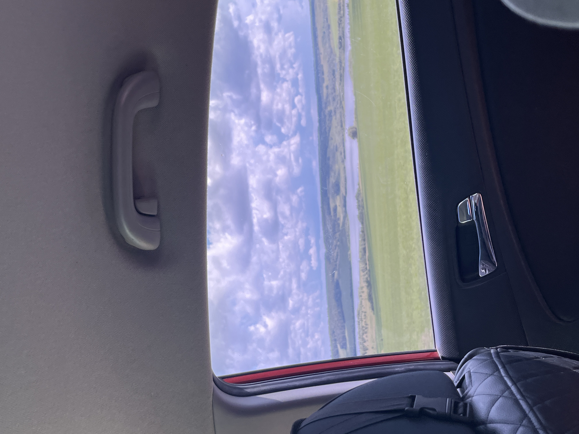 The image captures a view from the interior of a vehicle, looking out a window. Inside the car, the grey fabric of the headliner is visible at the top, with a plastic grab handle attached. Below this, a portion of a black quilted seat cover is seen on the left. The door panel, made of black plastic with a textured surface, is prominent in the lower right, featuring a silver door handle. Through the window, a landscape unfolds under a sky filled with white and grey clouds. The scene outside consists of rolling green hills, a body of water that appears to be a river or lake, and scattered trees. The overall impression is of a travel scene, possibly on a road trip, with the car interior framing the expansive natural scenery. The location in the city of Tolbazy, Russia, is not explicitly evident from the visual cues in the image alone.