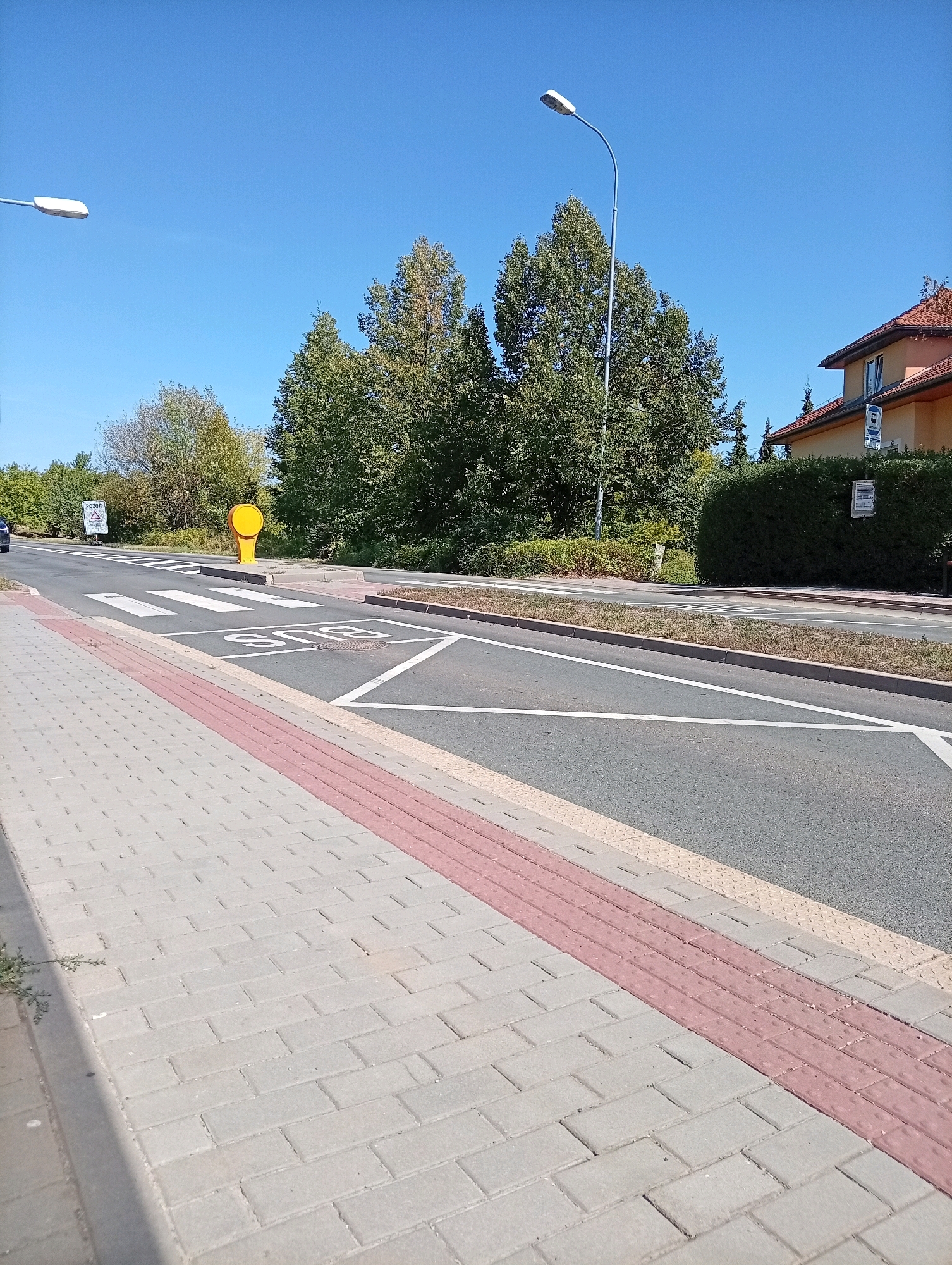 The image shows a street scene in Tišnov, Czechia. In the foreground, a gray brick sidewalk with a red tactile paving strip runs along the left side. The sidewalk leads towards a crosswalk painted with white stripes on the asphalt road. A yellow, cylindrical object is positioned next to the crosswalk, and a speed limit sign indicating "50" is visible on the road surface. A streetlamp with a white fixture arches over the road. Further down the road, trees and a building with an orange roof are visible, with a hedge in front of it. A bus stop sign is mounted on a pole near the building. The sky is clear and blue.