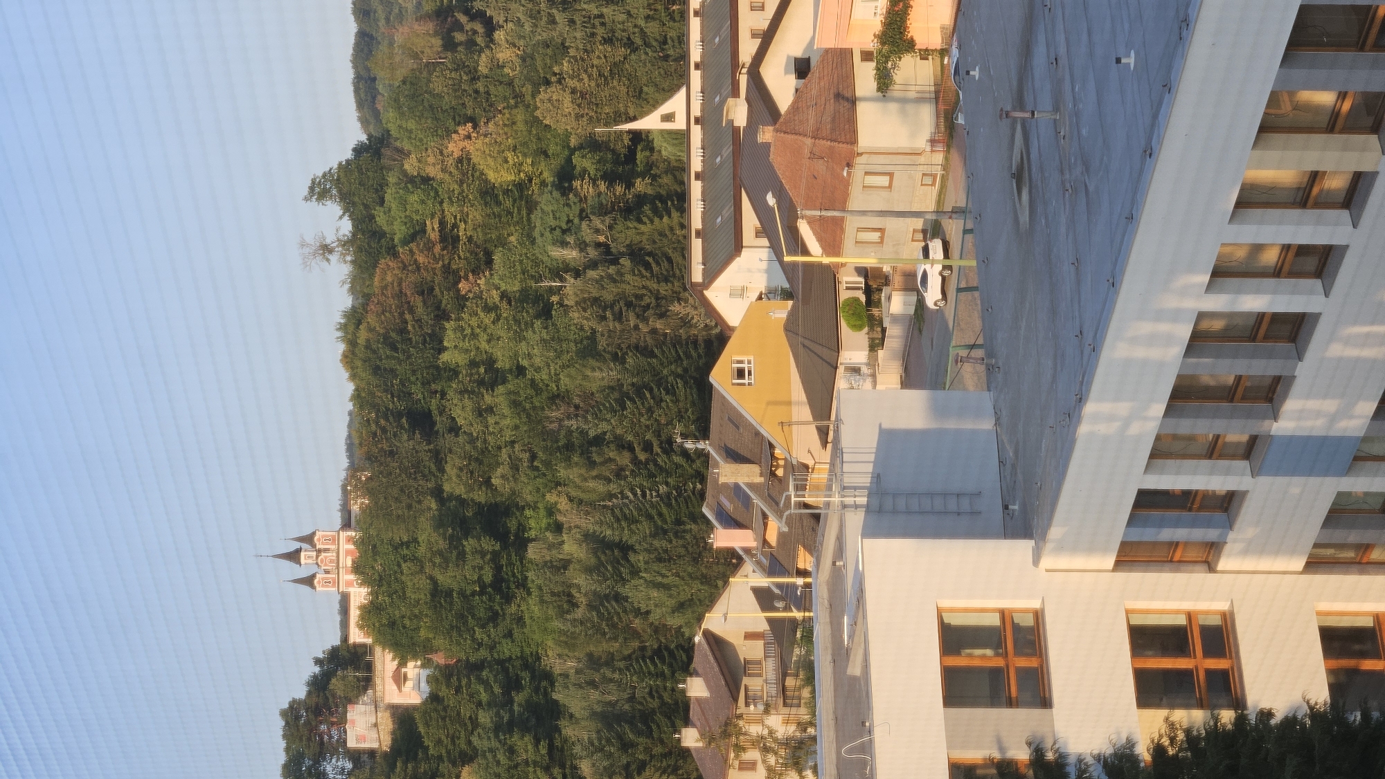 The image depicts a view of a residential area with a church visible in the distance on a forested hill. The foreground features modern multi-story buildings with flat roofs and numerous windows. The buildings are primarily white and grey, with some showing signs of wear or age.
In the middle ground, there are several houses with pitched roofs, some of which are tiled in red or brown. The houses vary in color, with yellow, beige, and white being prominent. Chimneys are visible on some of the roofs. Trees with green foliage are interspersed among the houses, and a street is visible with a couple of parked cars.
The background is dominated by a dense forest, with varying shades of green indicating different types of trees and possibly seasonal changes. On top of the hill, a distinctive church or similar religious building with multiple spires and a pink facade is visible. The sky is clear and a pale blue.
The image quality suggests it was taken from a window or balcony at an elevated position. The lighting indicates it is likely morning or late afternoon, with soft shadows cast across the scene. No people are visible in the image. The scene is tranquil and suggests a suburban or semi-urban environment.
Based on visual cues such as architectural styles and the general landscape, this scene is consistent with views found in the city of Prešov, Slovakia, specifically an area with residential housing nestled against a backdrop of hills and forests, with a prominent landmark like a church or monastery situated on elevated ground.