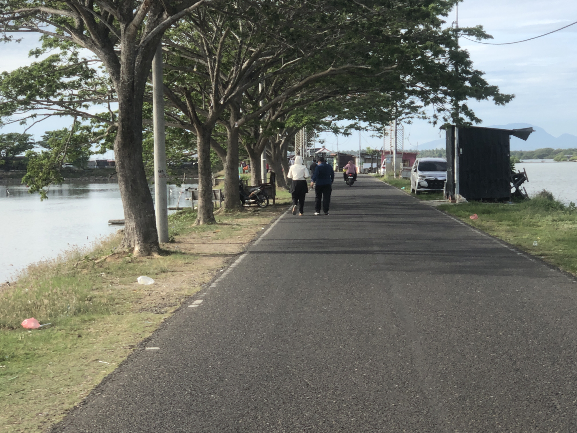 The image shows a road with a body of water on one side and trees lining the other. A couple walks down the road, away from the camera. A person on a motorbike is further down the road. A car is parked on the side of the road near a small structure. Trash is scattered on the grassy area near the water. The location is the city of Lhokseumawe, Indonesia.