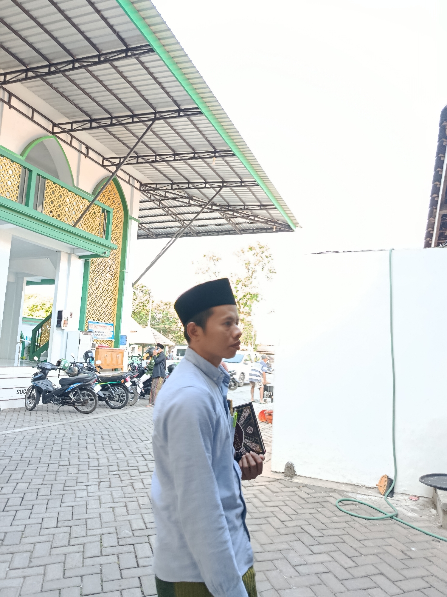The image depicts a man in a light blue long-sleeved shirt and a black cap, holding a book with a patterned cover, standing on a paved walkway outside a building with a green and gold facade. In the background, motorcycles are parked, and other people are visible. The building appears to be a mosque, suggested by its architecture and the presence of the book, which could be a Quran. The location is identified as Bondowoso, Indonesia. The sky is overcast. The scene is captured from a side-view angle, focusing on the man and the architectural details of the building.