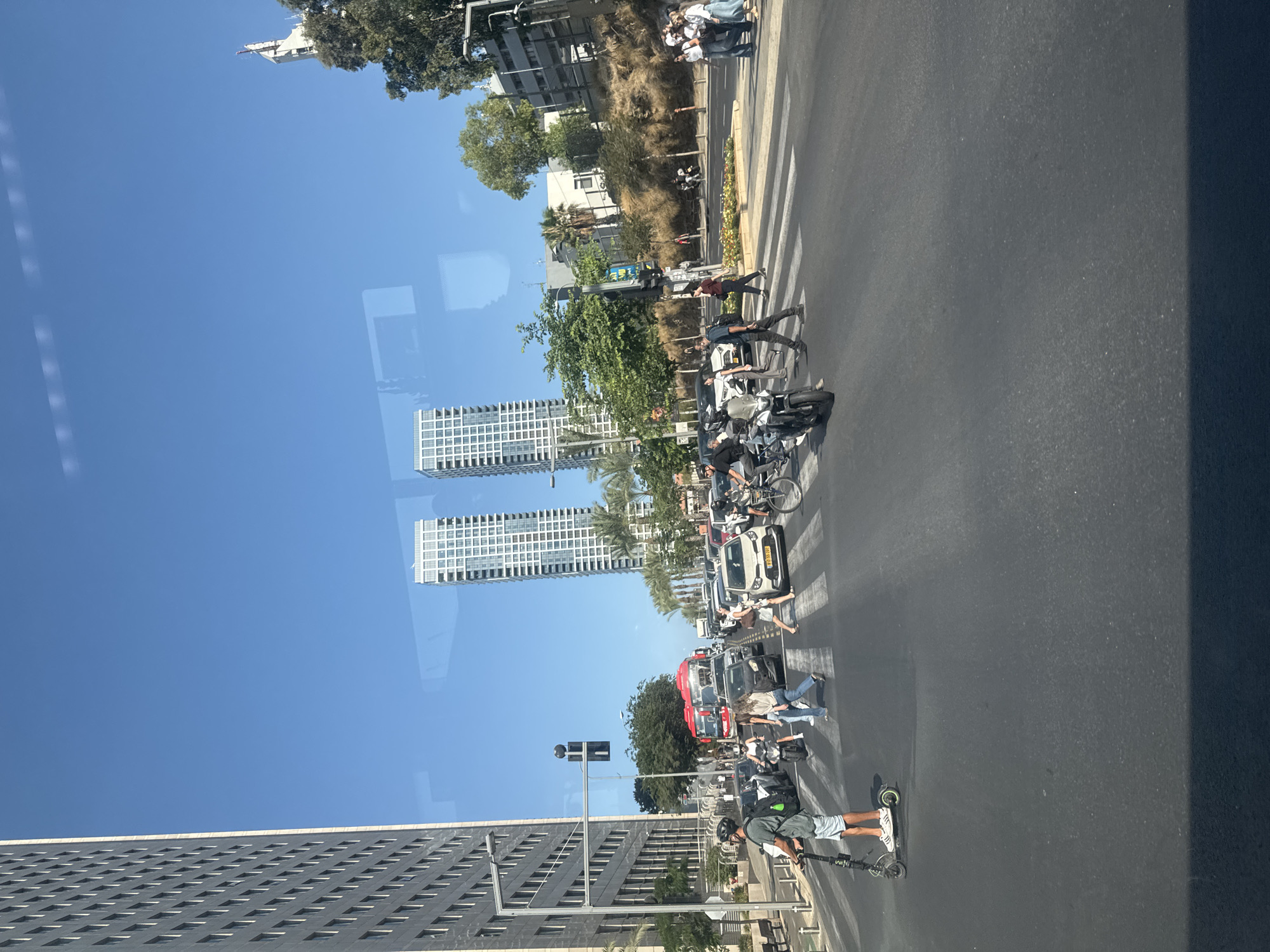 The image captures a street scene in Tel Aviv, Israel, under a clear blue sky. The foreground features an asphalt road with a pedestrian crossing. On the left side, a person is riding an electric scooter. Several cars and a motorcycle are present on the road, with some stopped at the crossing. Pedestrians are seen crossing the street. In the mid-ground, a red bus and other vehicles are part of a traffic jam. In the background, modern high-rise buildings and trees line the street. The overall impression is a busy urban environment on a sunny day.