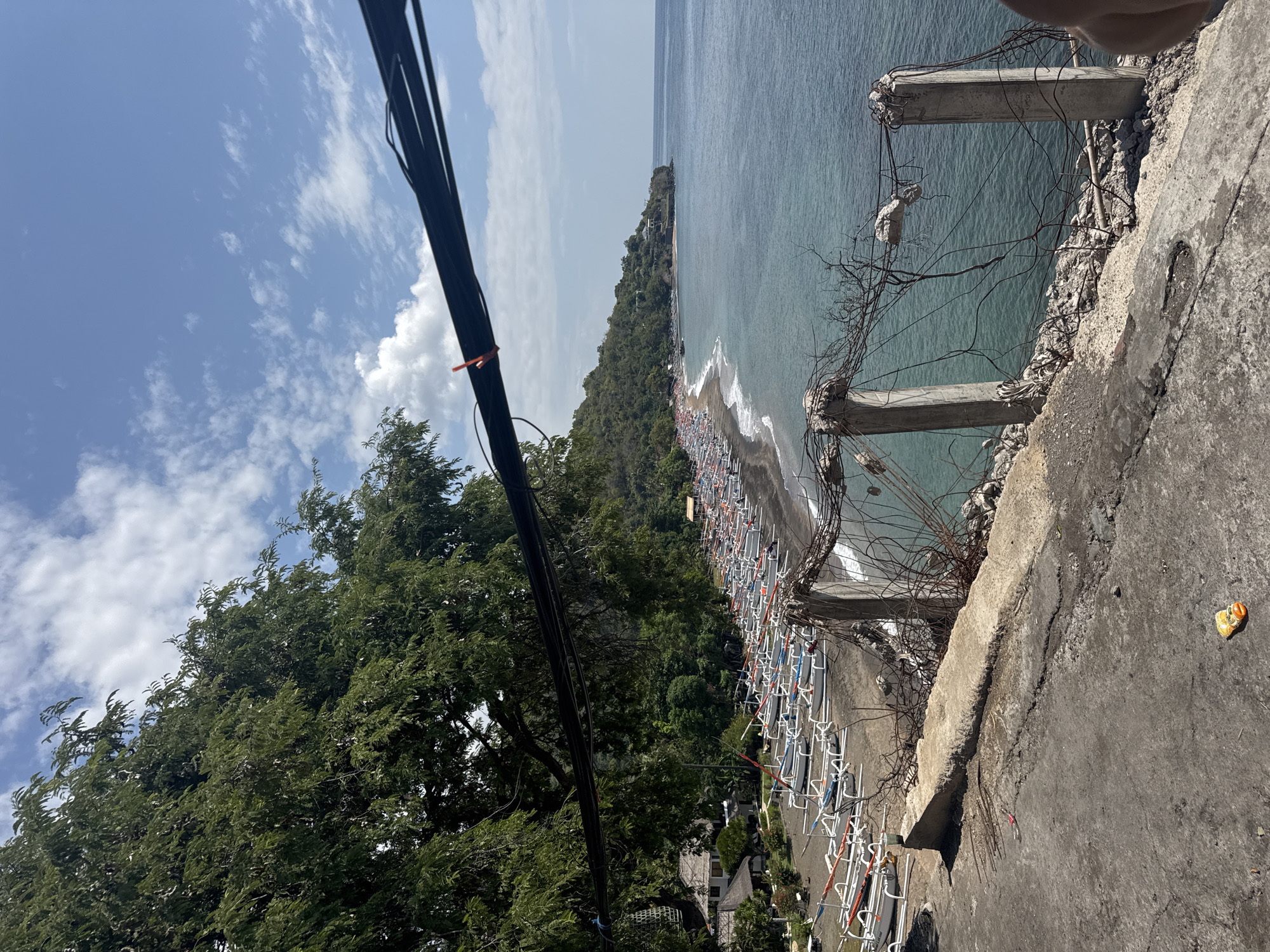 This is a medium-shot, eye-level photograph taken from a slightly elevated position overlooking a beach and the ocean. The scene is set in Abang, Indonesia, a coastal village known for its picturesque views.
In the foreground, a weathered concrete barrier runs along the edge of the frame, partially obscured by a dark, thick cable that stretches across the top of the image. The barrier is topped with rusted metal, tangled wires, and loose debris, suggesting a state of disrepair. A small, colorful object, possibly a piece of trash, lies on the concrete path in the lower left corner.
Beyond the barrier, a dark sand beach stretches into the distance, lined with a multitude of white and blue outrigger canoes. These canoes are neatly arranged, indicating they are likely used for fishing or local transportation. Further down the beach, a few buildings with thatched roofs are visible, nestled amongst lush green trees.
The ocean occupies the right side of the image, its water a clear, turquoise hue near the shore, transitioning to a deeper blue further out. The sea is calm, with small waves gently lapping at the beach. A long, green, tree-covered hillside rises from the coastline in the background, dotted with some scattered structures.
The sky above is a bright blue, with scattered white clouds adding texture to the scene. The overall impression is one of a tropical coastal landscape, with a mix of natural beauty and man-made elements, some of which appear neglected. The presence of the canoes and buildings suggests a local community that relies on the sea.