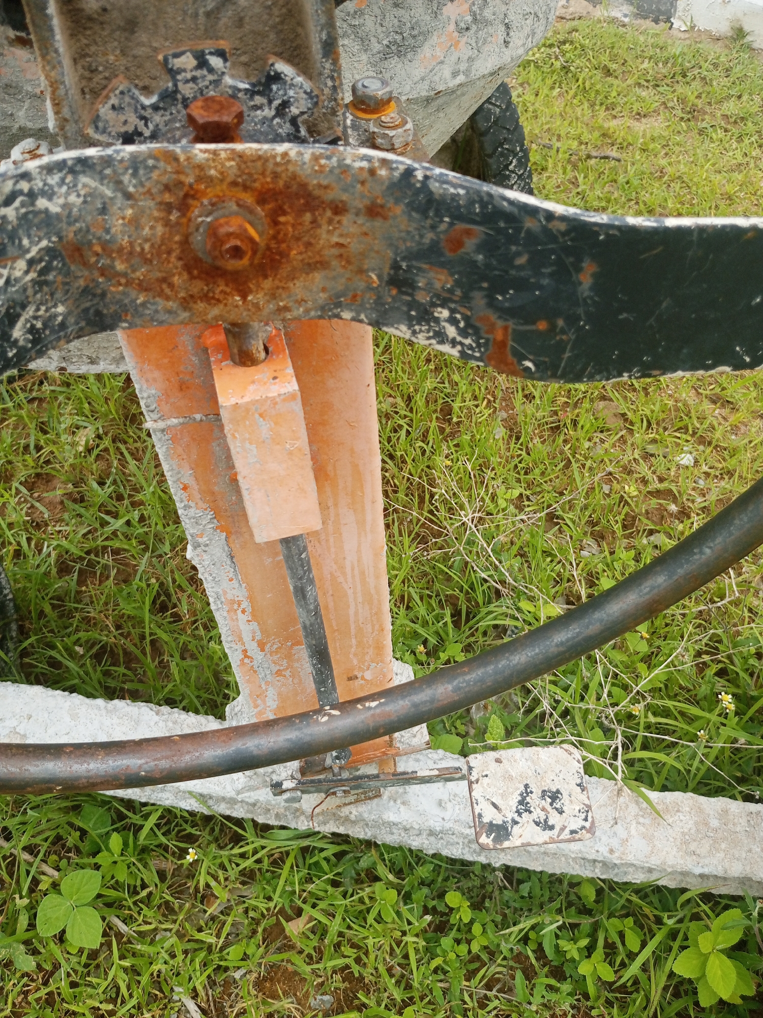 A close-up, eye-level shot captures a section of a rusted, orange and black metal structure, likely part of machinery, set against a background of green grass and soil. A prominent rusted bolt is visible on the black metal arm, with orange paint peeling away from the underlying metal. A dark, cylindrical metal pipe runs diagonally across the frame from the top right to the bottom left, resting on a rough, concrete-like surface. The concrete surface appears to be broken or uneven and is partially covered by grass. Small wildflowers, one white with yellow and another with yellow petals, are scattered in the grass. The scene suggests an outdoor environment, potentially in a rural or semi-rural area within Jalingo, Nigeria, given the vegetation and the presence of what could be construction or agricultural equipment.