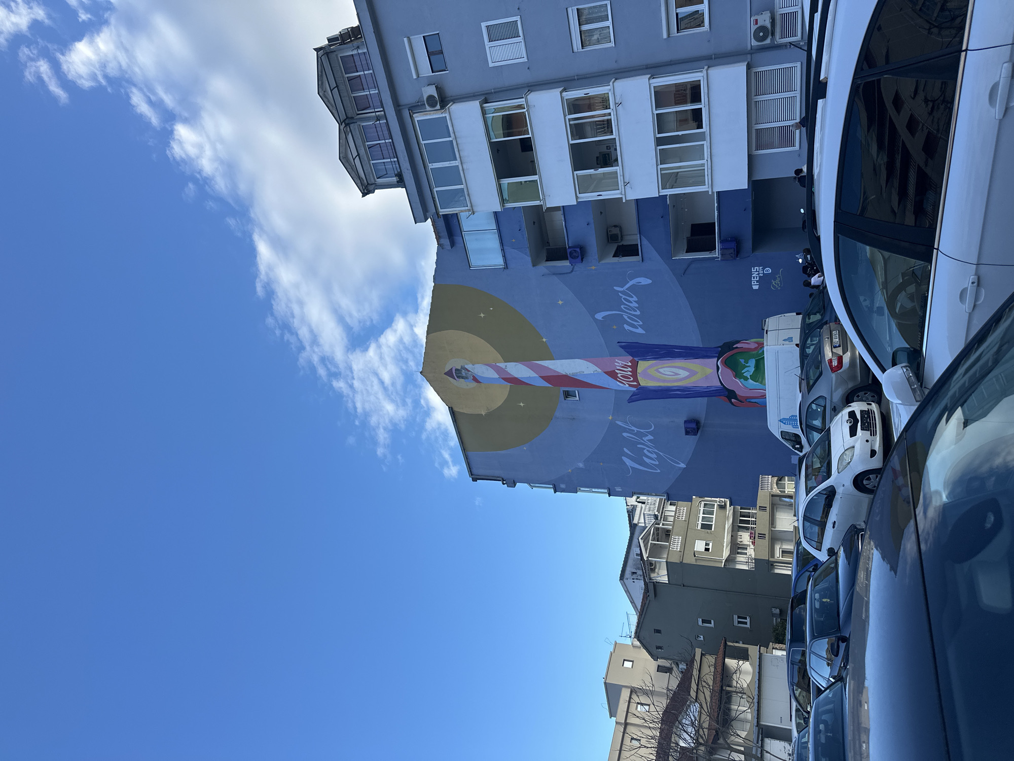 The image depicts a blue residential building in Budva, Montenegro, adorned with a mural of a stylized lightbulb. The mural features the words "light your ideas" written in a whimsical font. The building is partially obscured by parked cars, predominantly white, along the street. The sky above is a clear blue with scattered white clouds. Other buildings can be seen in the background.