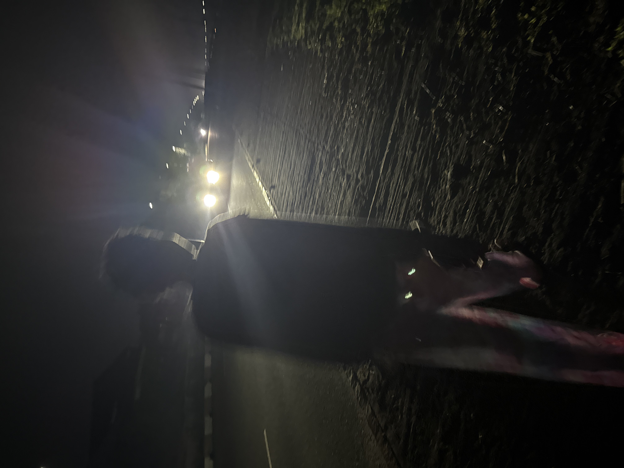 In Uyo, Nigeria, at night, a person stands on the shoulder of a road. The person, seen from behind, is blurred due to motion. They wear dark clothing on their upper body and colorful, possibly tie-dye, pants. Their silhouette is visible against the illuminated roadway, where the headlights of an approaching vehicle create bright streaks of light. The road is paved and marked with white lines. To the right of the road, there is an area of grass and vegetation. The background is dark, with other distant lights visible along the road. The overall scene conveys a sense of movement and a nocturnal urban environment.