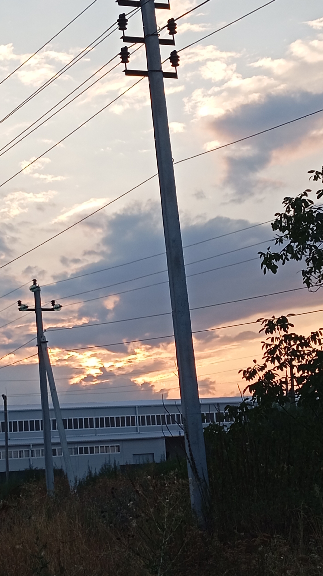 The image shows a power pole with wires in the foreground against a sunset sky. In the background, a long, modern building with many windows is visible. There is also vegetation in the foreground and right side of the image. The location is identified as Baksan, Russia.
