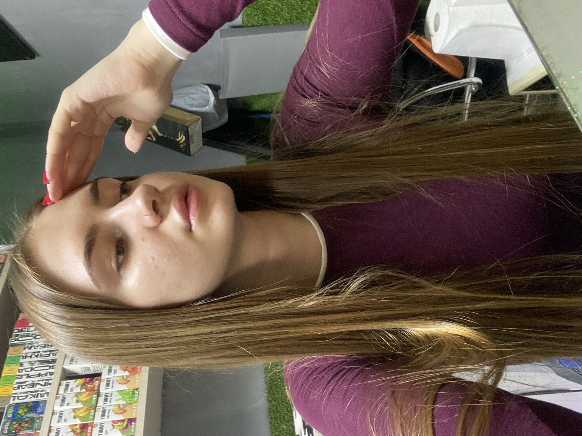A young woman with long, straight, light brown hair is shown in a medium shot, seated indoors. She is wearing a dark magenta long-sleeved shirt with a white trim around the neckline. Her right hand is raised to her forehead, with her fingers slightly splayed, and her fingernails are painted red. She has a subtle nose ring. The background reveals shelves stocked with various items, including what appear to be boxes of vape liquids and other products, suggesting a retail environment. A roll of white paper towels is visible on a desk or counter in the foreground to the right. The lighting suggests an indoor setting, possibly a shop or office. The image's metadata does not contain any location information for Дьяково, Russia.