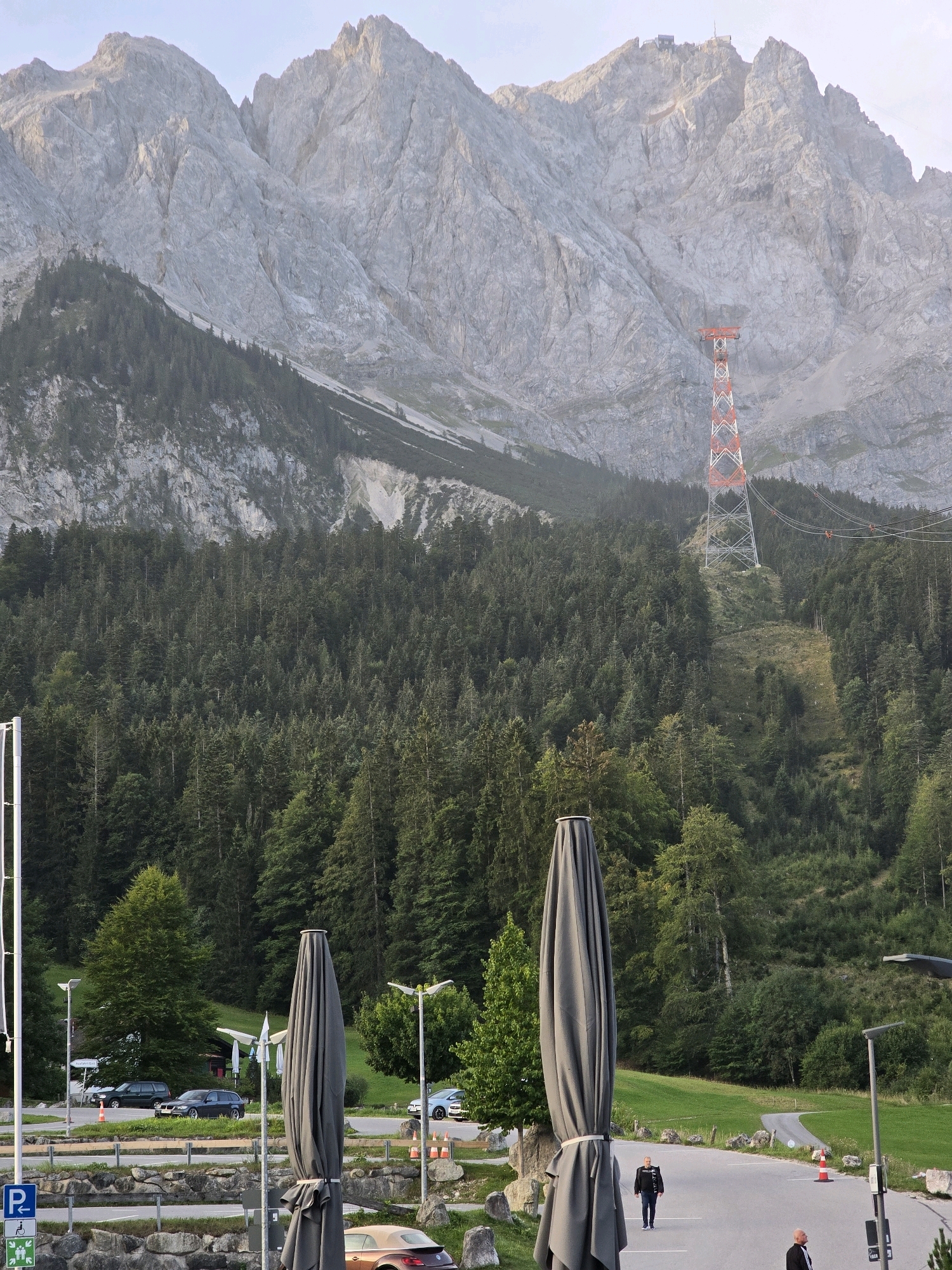 This is a photograph of the Zugspitze in Grainau, Germany. The image captures a vast mountainous landscape with a prominent cable car tower in the mid-ground, supporting a cable that stretches across the scene. The tower is red and white, a striking contrast against the muted tones of the mountains and the lush green forest that blankets the lower slopes. The mountains themselves are rugged and imposing, with visible rock faces and crevices.
In the foreground, there's a paved area that appears to be a parking lot. Several cars are parked here, including a dark sedan, a light blue sedan, and a partially visible orange convertible. Two large, closed umbrellas, likely for outdoor seating, are prominent in the foreground, obscuring some of the view.
Two men are visible on the paved surface. One man, wearing a black jacket and dark pants, stands facing the camera. Another man, also in dark clothing, is further in the background, walking away from the camera. The environment suggests a recreational area at the base of the mountain, possibly a viewpoint or a station for the cable car. The sky is a pale, clear blue, indicating fair weather.