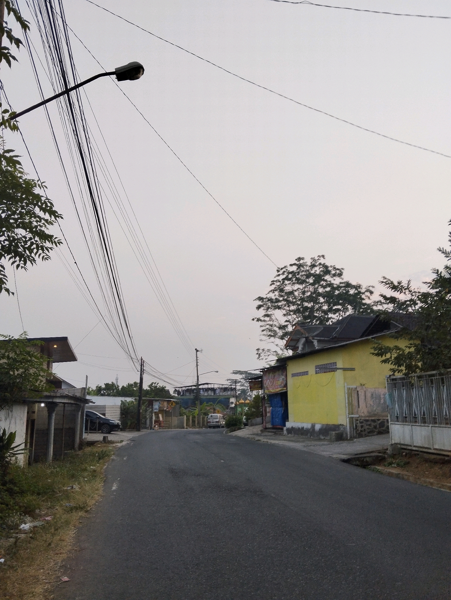 The image depicts a road in Salatiga, Indonesia, with buildings and overhead wires lining the street. The sky is overcast, suggesting it might be dusk or a cloudy day. On the left side of the road, there is a building with a white facade and pillars, partially covered by trees. Further down the road on the left, there is another structure with a dark roof and a yellow facade. On the right side of the road, there is a yellow building with a dark roof and a fence in front. There are several cars visible on the road and parked near the buildings, including a dark-colored sedan and a light-colored van. Numerous overhead wires crisscross the sky, supported by poles. The overall impression is of a typical urban or semi-urban street scene in Indonesia.