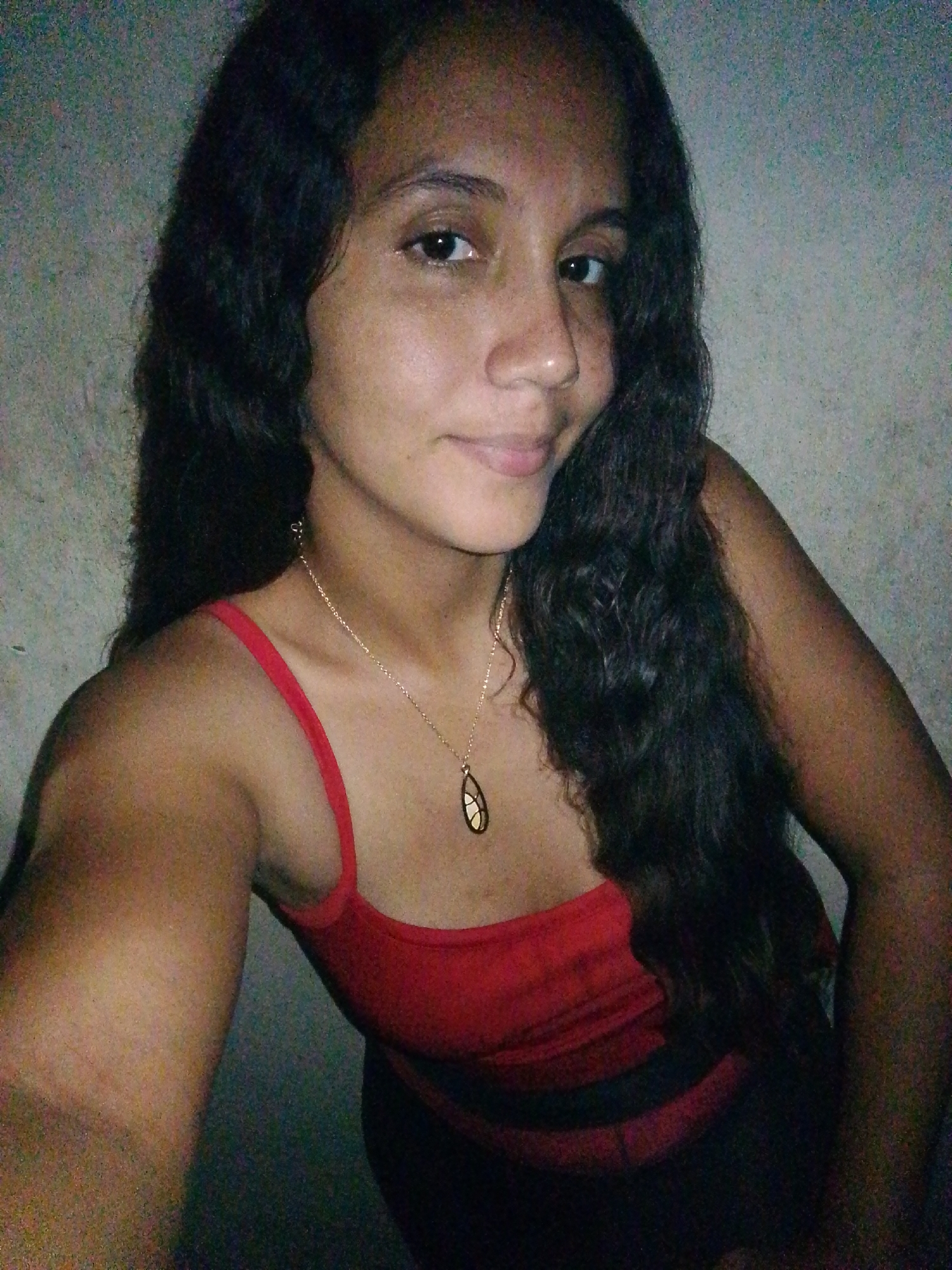 A young woman with long, dark, wavy hair is posing for a selfie indoors against a light-colored wall. She has dark eyes, a medium complexion, and is wearing a red tank top with black trim. She is also wearing a gold necklace with a teardrop-shaped pendant. The location is in the city of Juan Griego, Venezuela.