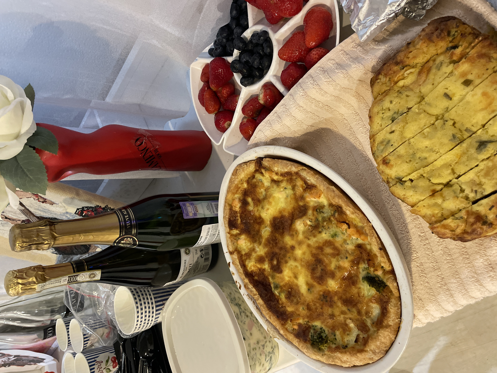 The image depicts a still life arrangement on a table. There is a white floral arrangement in the upper left corner. Next to it is a red container and a white sectioned dish filled with strawberries and blueberries. Two bottles of sparkling wine are visible, lying horizontally across the table. Cups and white containers are grouped near the wine bottles. A quiche in a white oval dish sits on a light-colored towel, next to a sliced frittata.
The city of Sochi, Russia is not discernible from the image content.