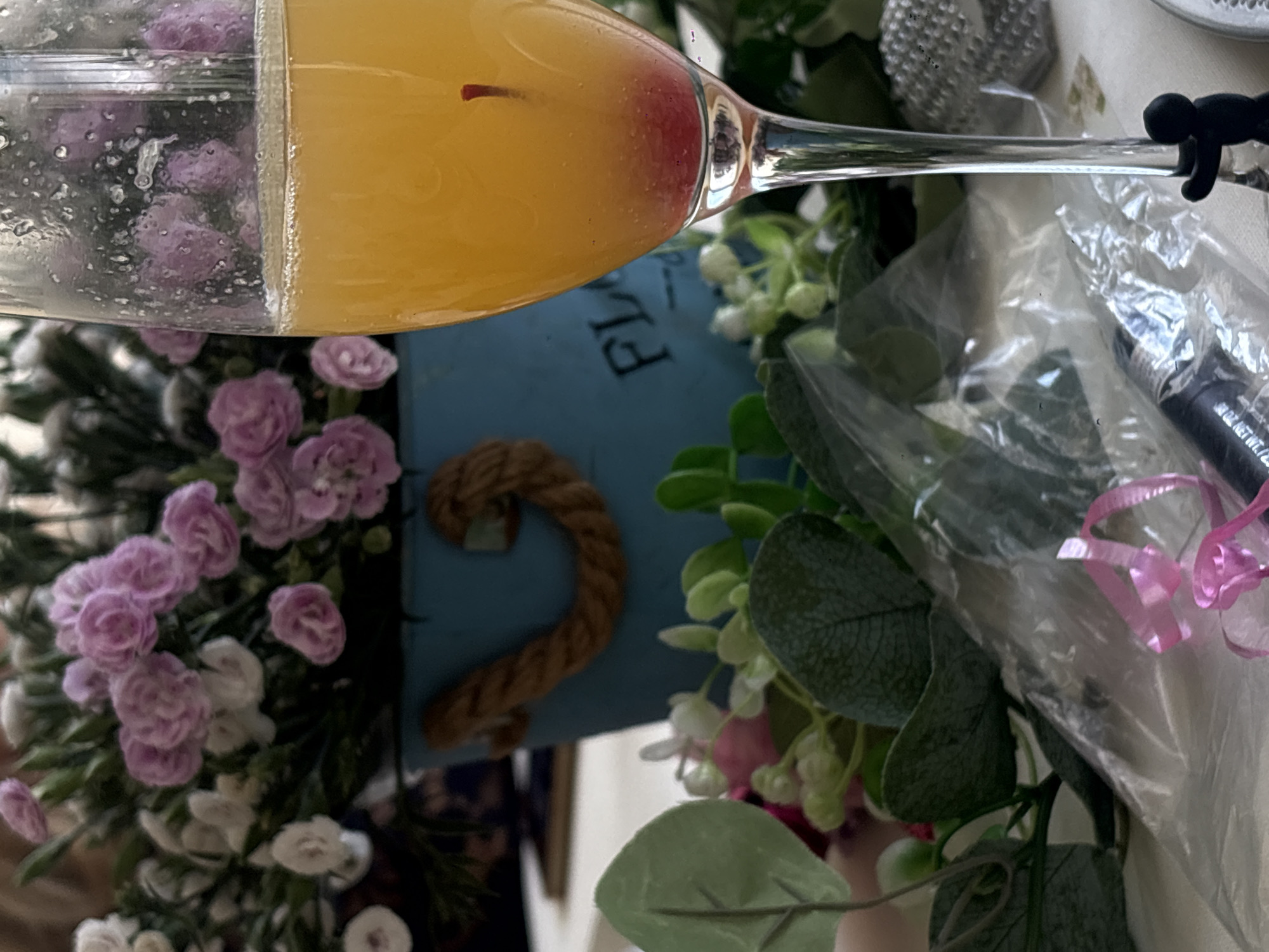 The image shows a close-up of a champagne flute filled with an orange liquid and a single red berry. The flute rests on a white tablecloth. In the background, a blue bucket with rope handles contains a bouquet of pink and white artificial flowers. To the left and right of the flute, there are more artificial green leaves and pink flowers. In the foreground, a clear plastic wrapper with pink ribbon and a black tube are visible. A small black figurine is clinging to the stem of the champagne flute. The scene appears to be indoors or in a sheltered outdoor setting, given the artificial flowers and tablecloth. The location is not discernible from the image itself, and no information about Santiago Yancuitlalpan, Mexico, can be inferred.