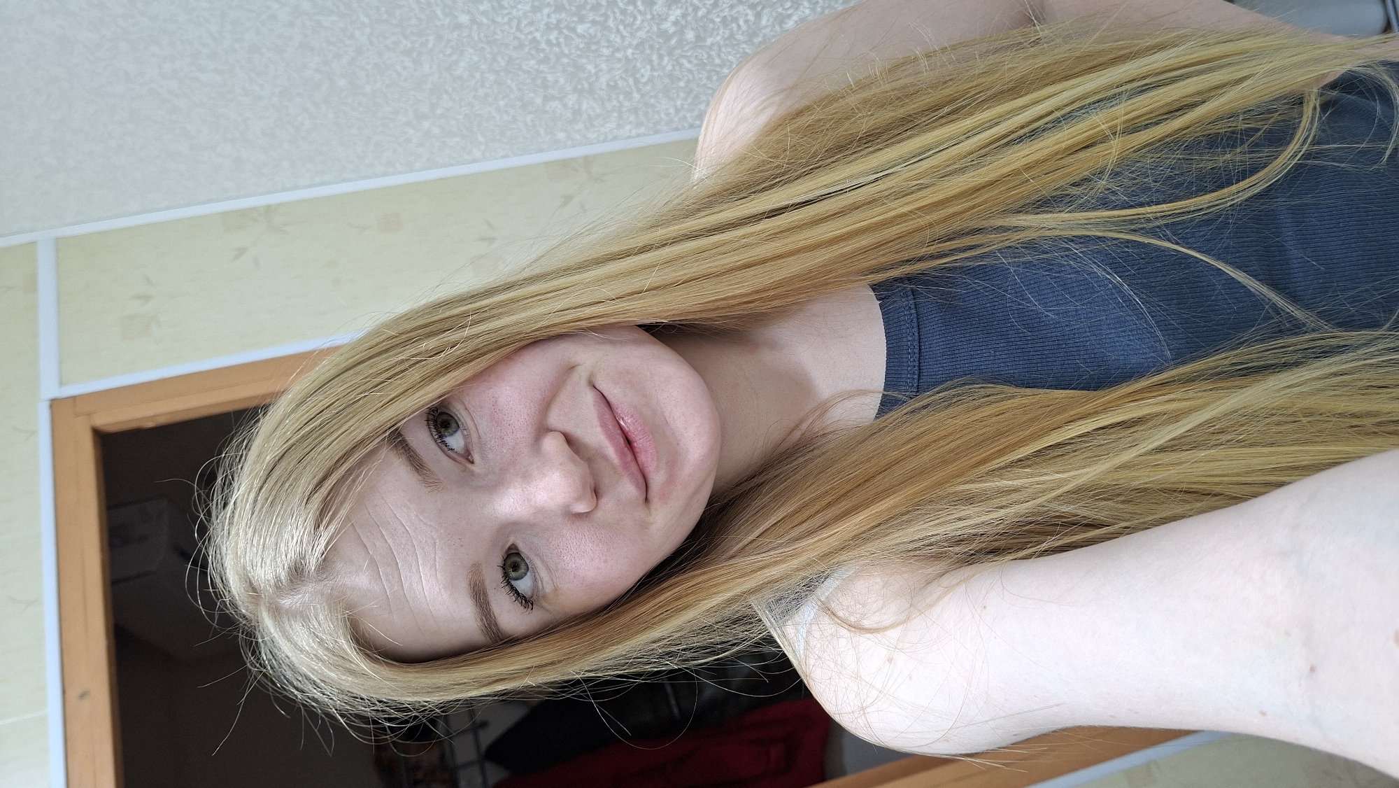 A fair-skinned woman with long blonde hair is wearing a grey tank top in what appears to be an indoor setting with yellow wallpaper and a brown door frame. The image is taken from a slightly tilted perspective. The location is Большой Букор, Russia.