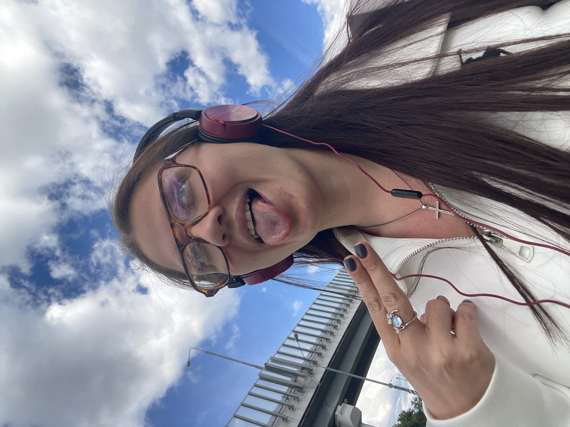 A young woman is pictured in an upward-facing selfie. She is wearing glasses, headphones, a white jacket, and a silver necklace with a cross pendant. Her hair is long and brown. She is sticking her tongue out and displaying a ring on her finger. The background consists of a blue sky with white clouds, a bridge, and a few light poles. Given the location information, the image was taken near Razvilka, Russia.