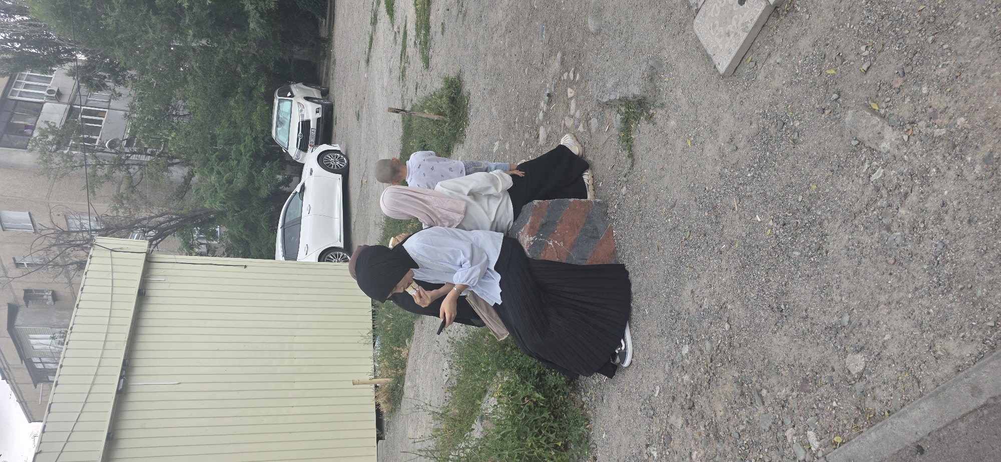 In an outdoor urban setting, likely in Bishkek City, Kyrgyzstan, three individuals are present. Two women, dressed in hijabs and long skirts, are seated on a low, segmented concrete barrier. The woman on the left, wearing a black hijab and a light blue shirt, is holding a mobile phone and appears to be eating. The woman to her right wears a light-colored hijab and a white top. A young boy, facing away from the camera, stands near the women. In the background, two white cars are parked, and a multi-story apartment building is visible, along with trees and vegetation. The ground is covered with gravel and dirt.