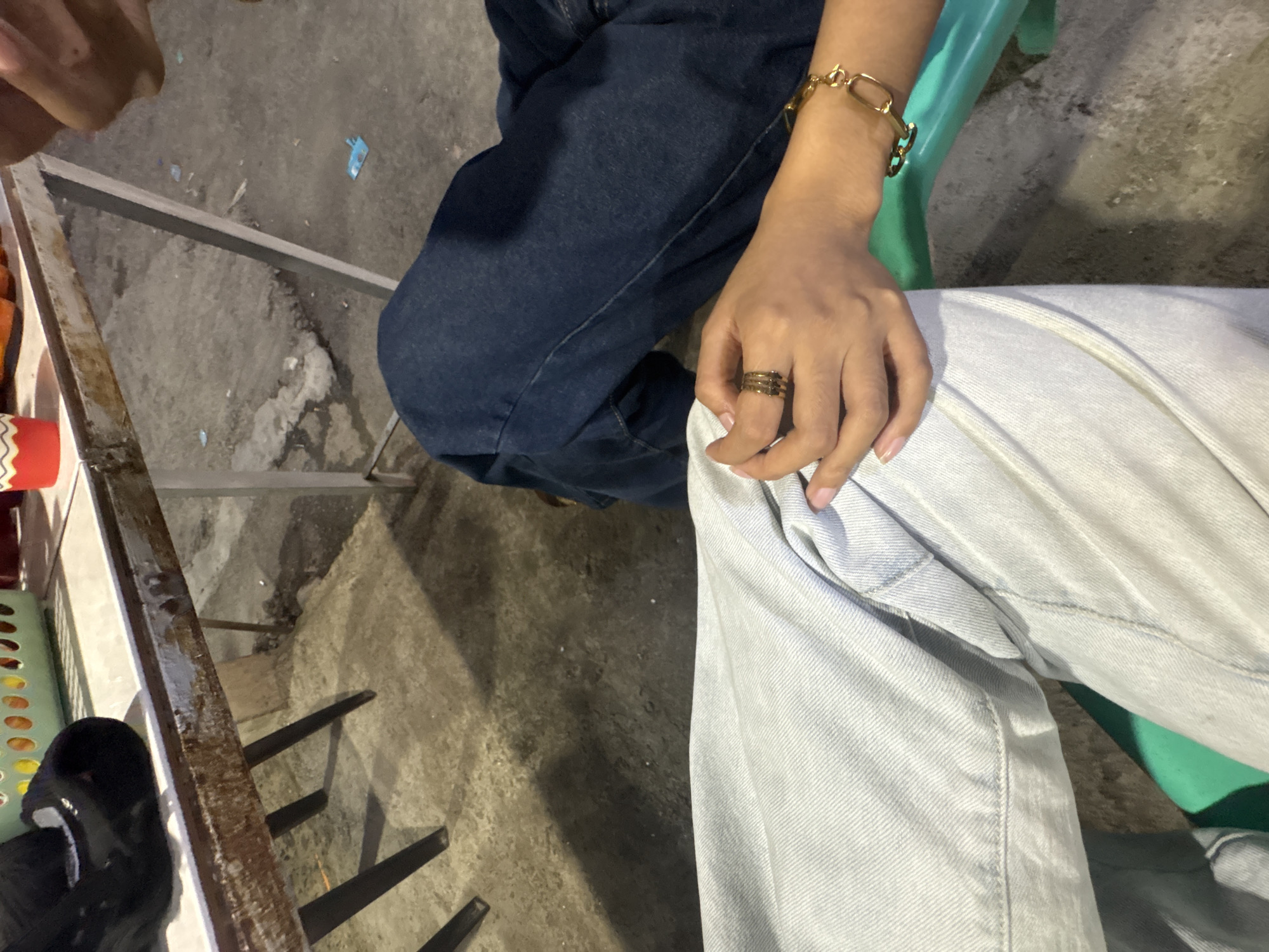 The image depicts a close-up view of two people sitting on a plastic chair next to a metal table. The person on the left is wearing light blue denim jeans. The person on the right is wearing dark blue denim jeans and has a gold bracelet on their left wrist, as well as a gold ring with multiple bands on their left index finger. The metal table has a worn surface and appears to be outdoors, with a concrete ground visible beneath. A light-colored basket with circular items is visible on the table, along with a red and white cup. The scene suggests a casual setting, potentially a street food stall or a small eatery in Carmen, Philippines, given the context provided.