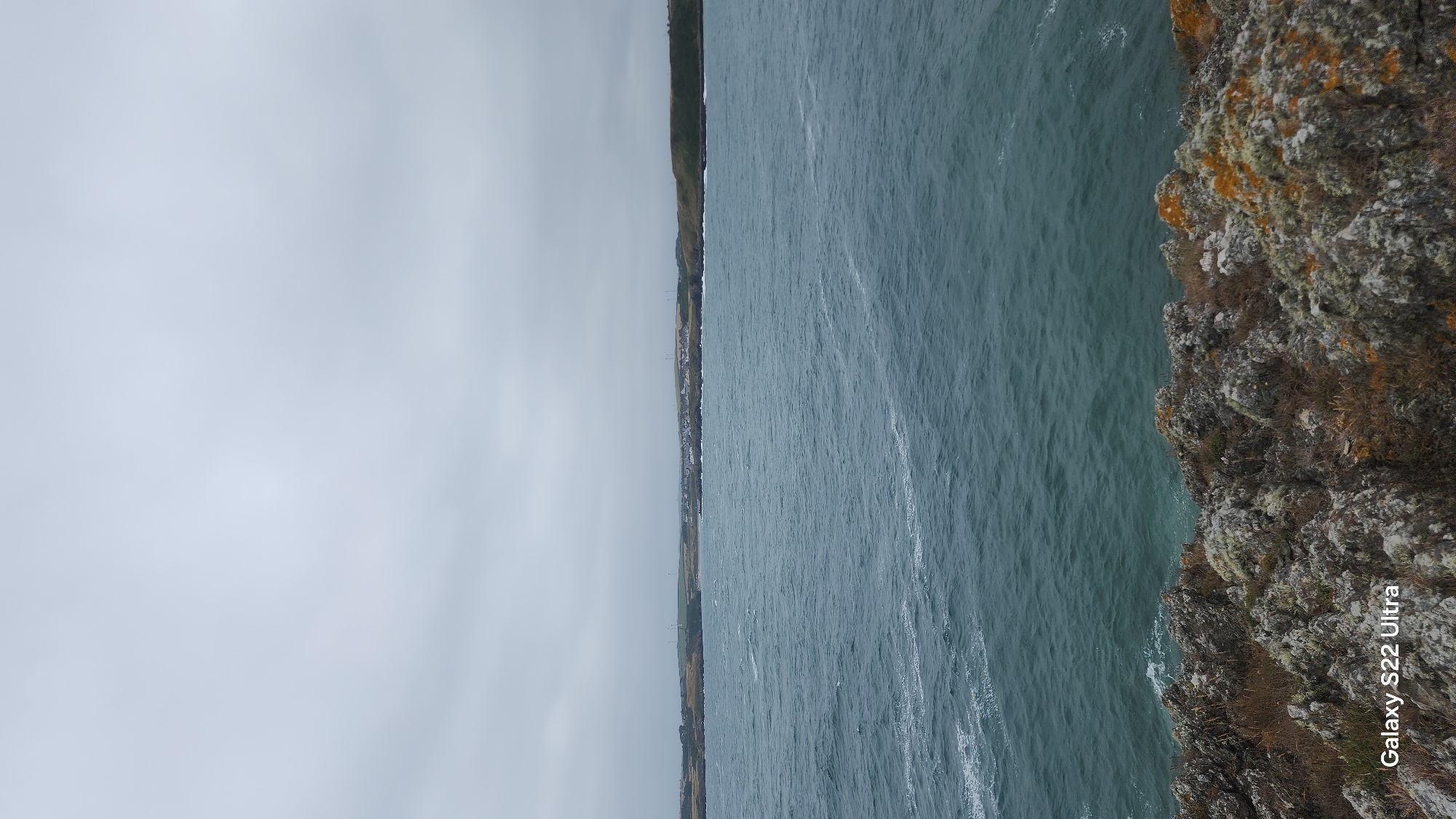 The image depicts a choppy, dark blue sea under a heavily overcast sky. In the distance, a low-lying coastline is visible, dotted with houses and some wind turbines. The foreground is dominated by a rugged, rocky cliff face covered in lichen and dry vegetation. The scene appears to be located on the coast of Cemaes, United Kingdom, given the presence of the "Galaxy S22 Ultra" watermark which is associated with mobile phone imagery. No people or specific actions are visible in the image.