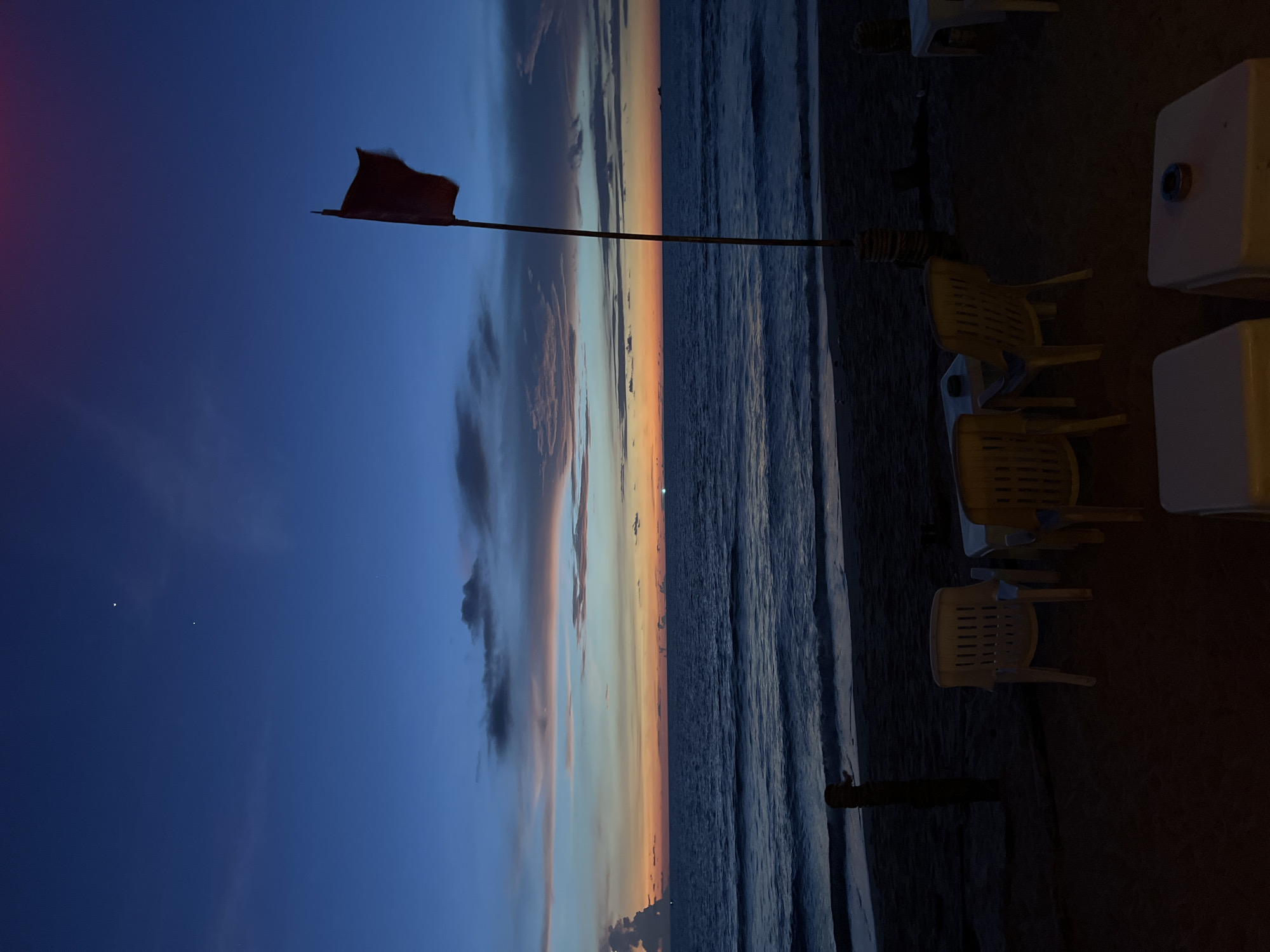 At dusk, a beach scene unfolds with a twilight sky of deep blues and oranges. The ocean, dark and undulating, meets the sandy shore. On the beach, several white plastic chairs and tables are arranged, some with ashtrays on them, suggesting a seating area for patrons. A tall flagpole stands near the water, bearing a red flag, which might indicate a warning or a specific zone. Two bright celestial bodies, possibly stars or planets, are visible in the upper portion of the sky. The overall atmosphere is serene, capturing the transition from day to night on a tranquil beach.