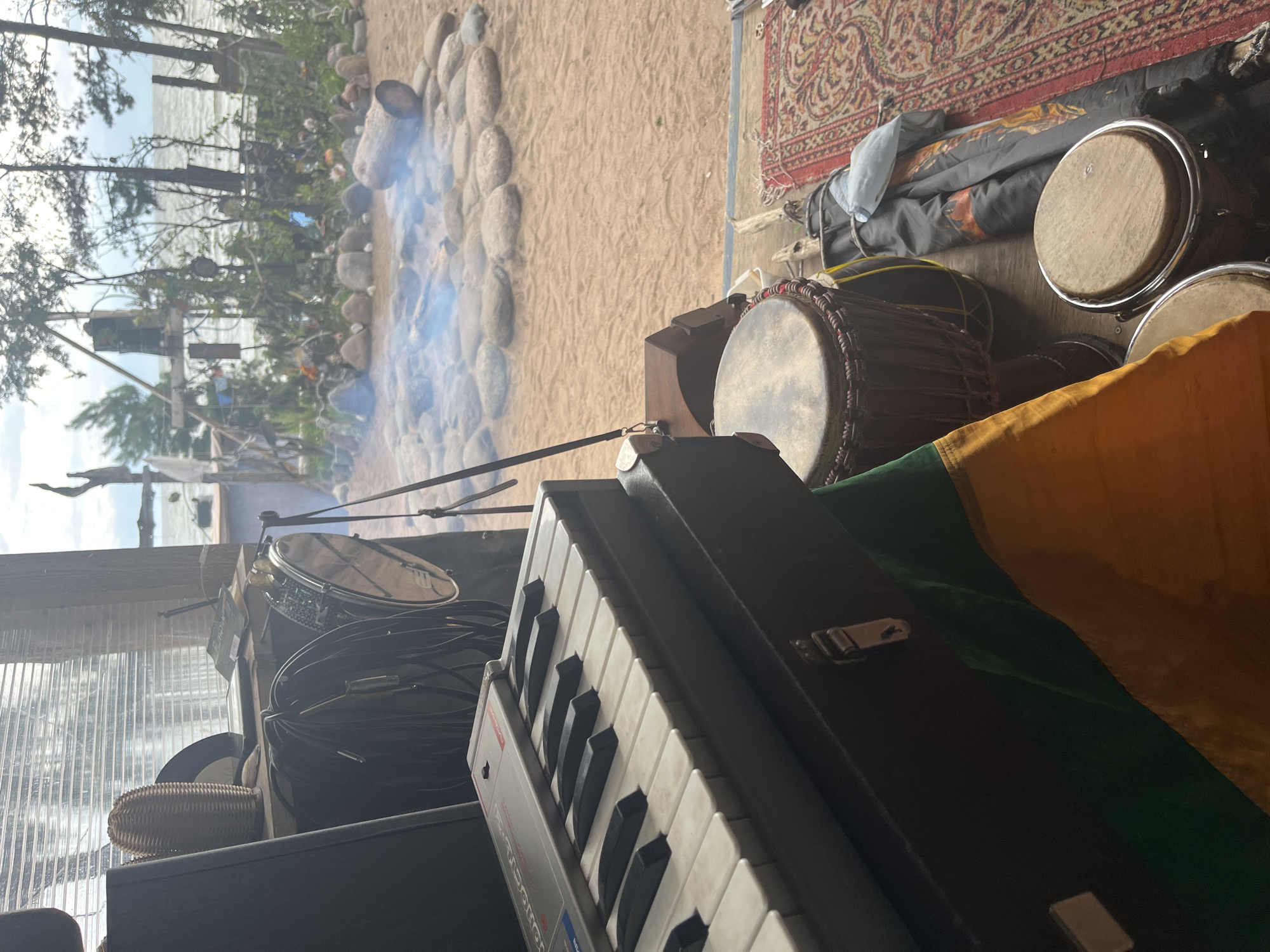 An eye-level, medium shot captures a collection of musical instruments and a cozy outdoor setting. In the foreground, a portable keyboard sits atop a dark case, partially covered by a green and yellow fabric. To the right, two drums, one larger and darker than the other, rest on a wooden floor. A red and gold patterned rug is laid out nearby. Beyond these objects, a sandy area is visible, featuring a stone fire pit with a log and wisps of smoke rising. A body of water stretches into the distance, lined by trees and a rustic wooden structure. The overall ambiance suggests a relaxed, perhaps bohemian, atmosphere.